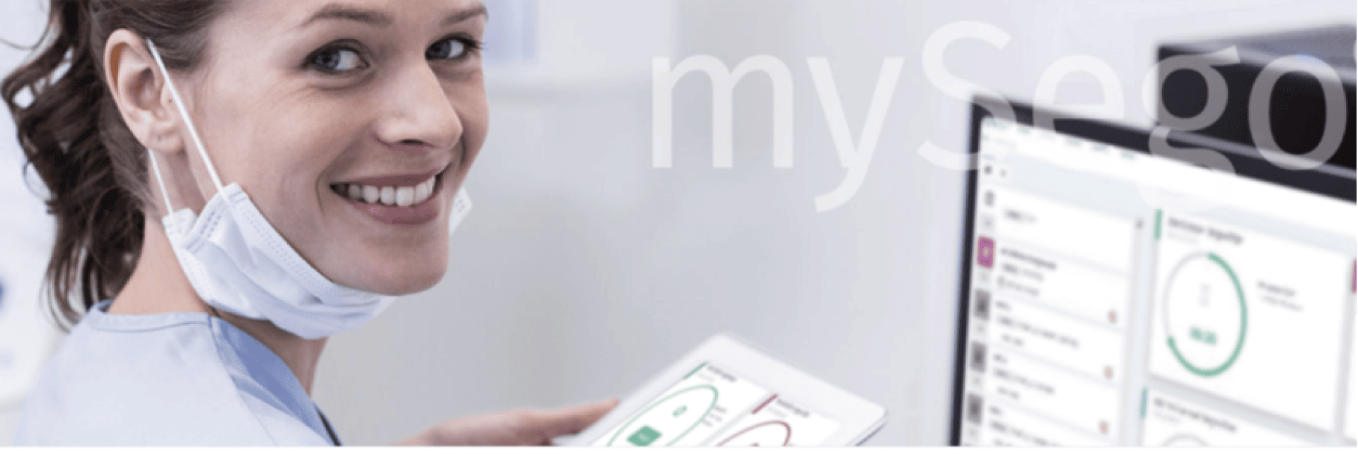 Une femme portant un masque sous le menton sourit à la caméra tout en tenant une tablette affichant des graphiques, avec un écran d'ordinateur affichant des données similaires en arrière-plan.