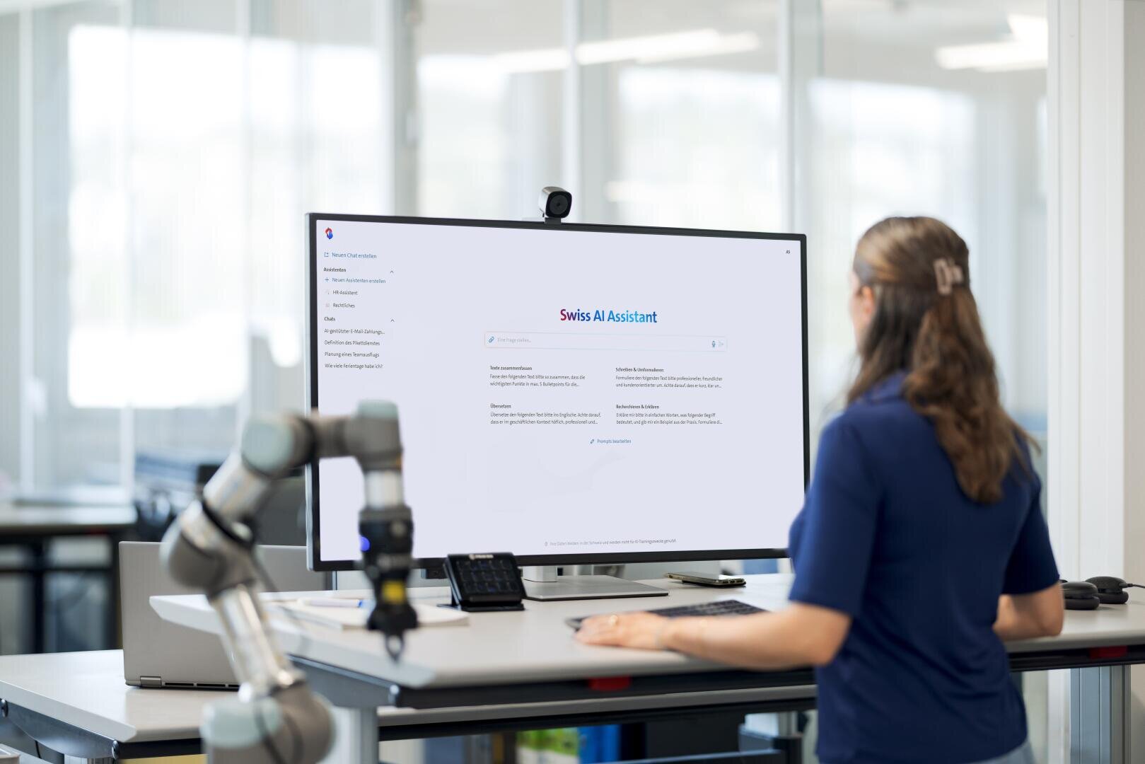 Eine Person steht an einem Schreibtisch mit einem großen Monitor, auf dem die Oberfläche des Swiss AI Assistant angezeigt wird. Bürogeräte und ein Roboterarm sind in einem hellen, modernen Arbeitsbereich zu sehen.