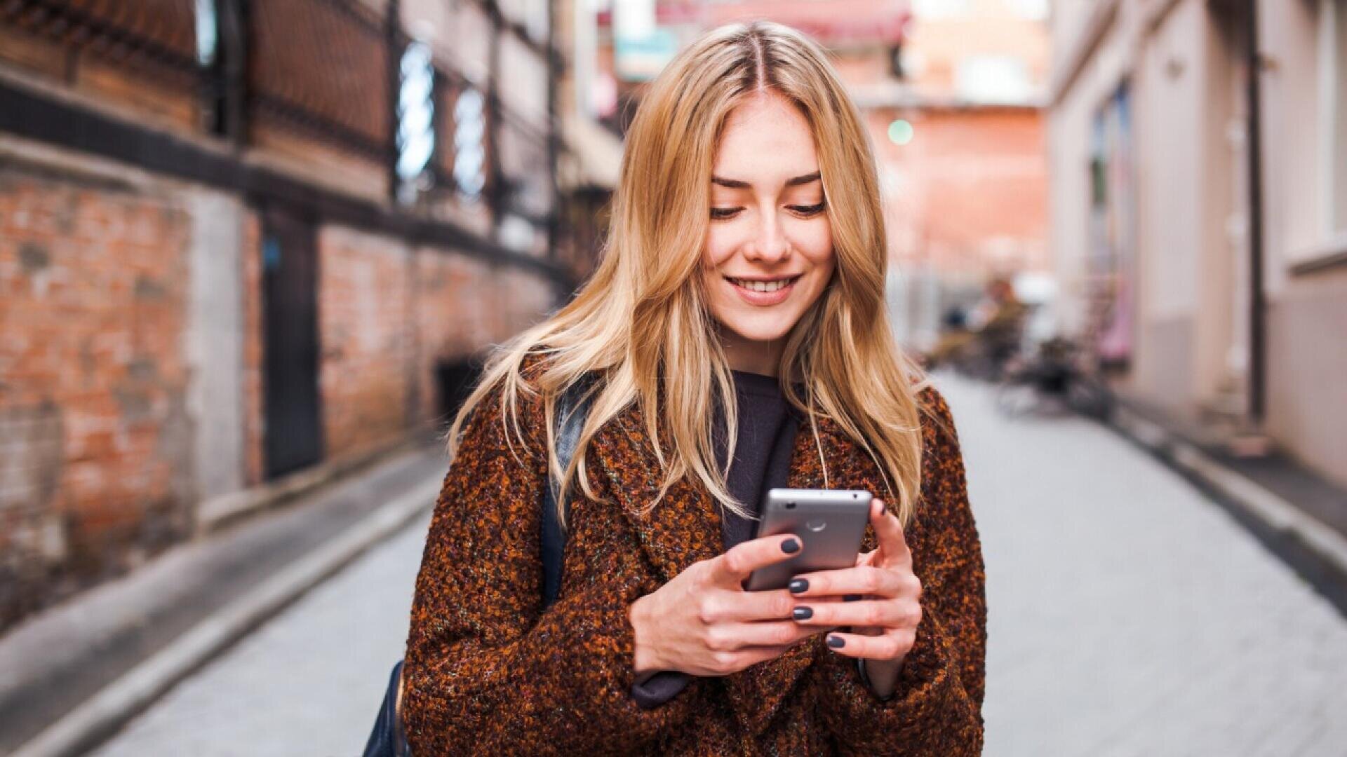 Une jeune femme aux longs cheveux blonds sourit en regardant son smartphone, debout en plein air dans une rue étroite avec des murs en briques et des bâtiments en arrière-plan. Elle porte un manteau marron et un sac à dos.