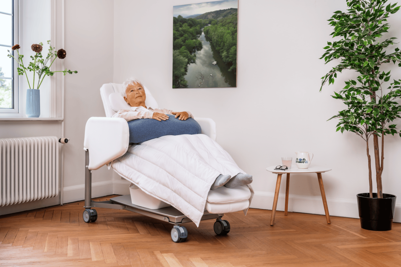 Eine ältere Frau macht es sich mit einer Decke in einem Liegesessel gemütlich – in einem hellen Raum mit Pflanzen, Blumen und einem Landschaftsgemälde an der Wand.