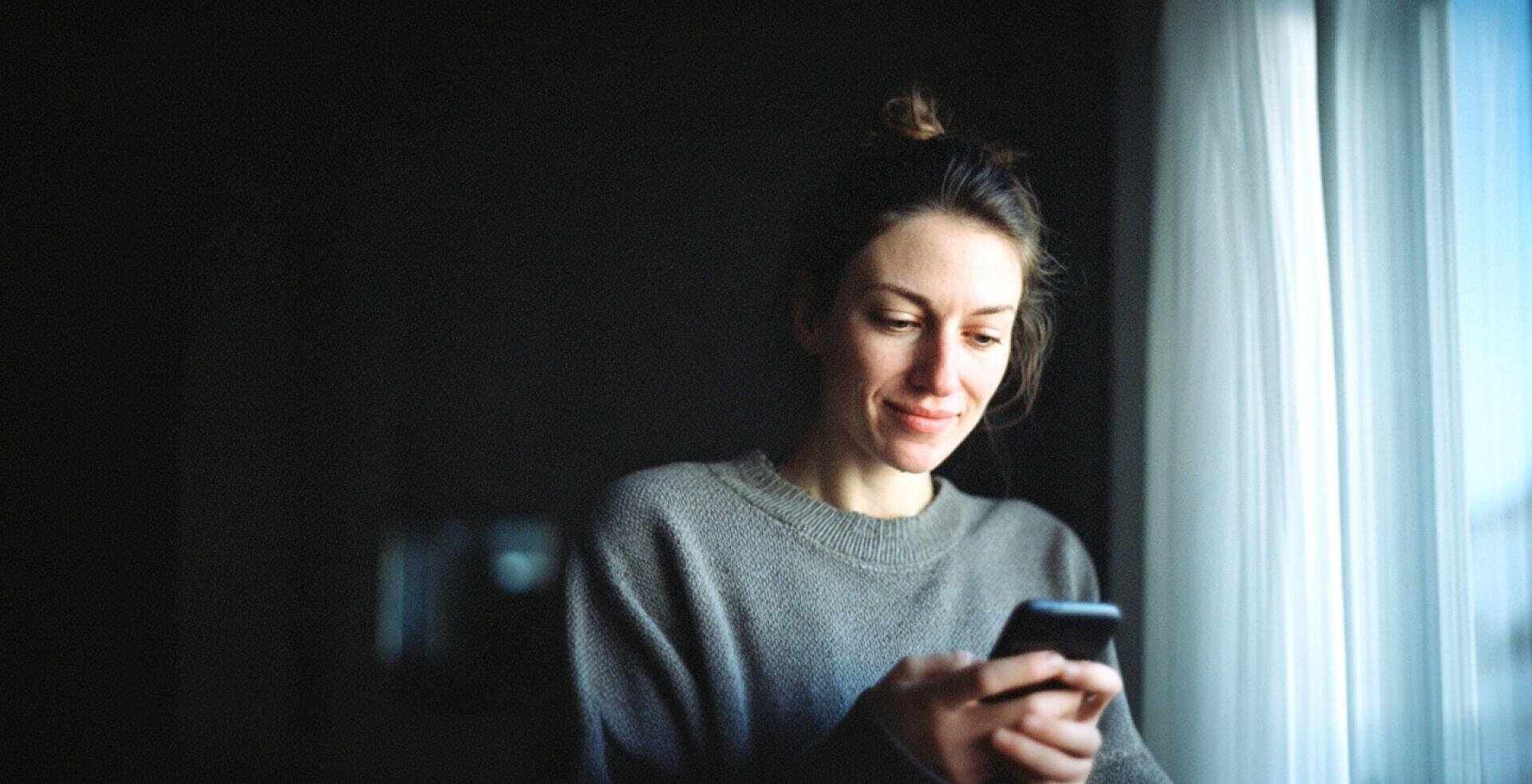 Eine Frau in einem grauen Pullover steht neben einem Fenster mit durchsichtigen weißen Vorhängen und lächelt, während sie auf ihr Smartphone schaut.