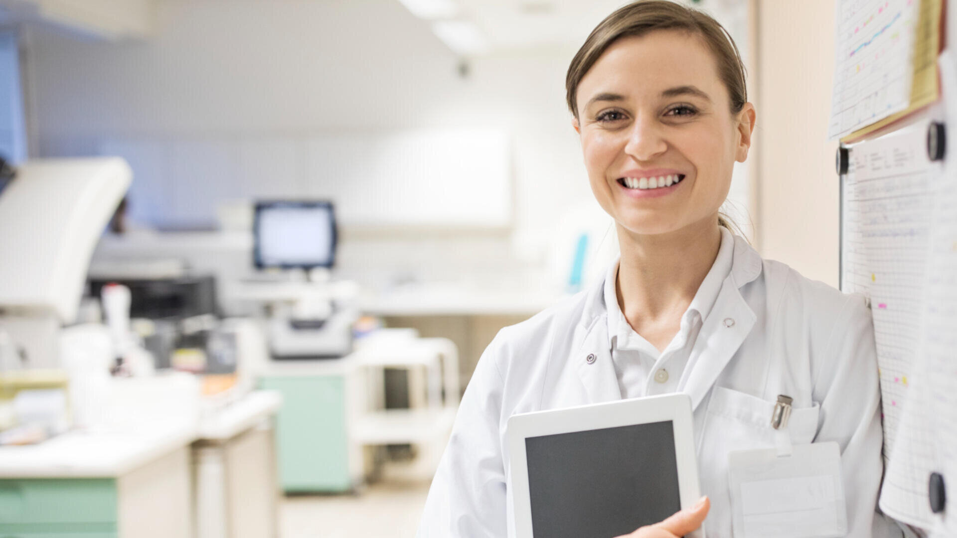Eine lächelnde Ärztin in einem weißen Laborkittel steht in einer Arztpraxis, hält ein Tablet in der Hand und blickt in die Kamera, während im Hintergrund medizinische Geräte und Krankenblätter zu sehen sind.