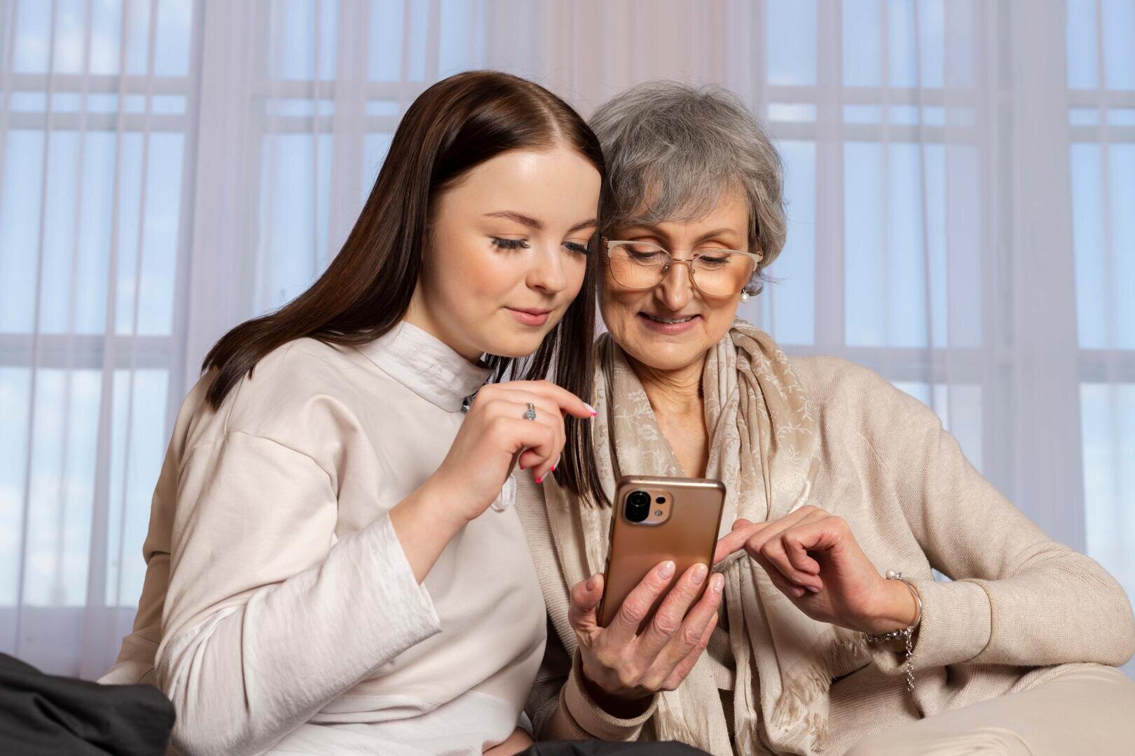 Une jeune femme et une femme plus âgée sont assises côte à côte sur un canapé ; elles sourient et regardent un smartphone que la femme plus âgée tient à la main. La lumière pénètre à flots par de grandes fenêtres, derrière lesquelles se trouvent des rideaux vaporeux.