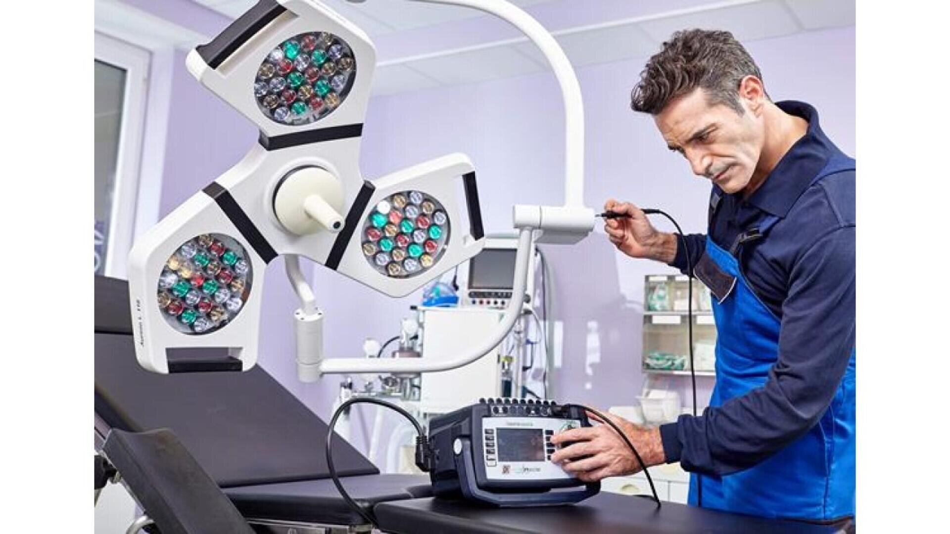 Un technicien en vêtements de travail bleus teste une lampe chirurgicale à l'aide d'un équipement électronique dans une salle d'examen médical moderne, à côté d'une table d'examen et d'appareils médicaux.