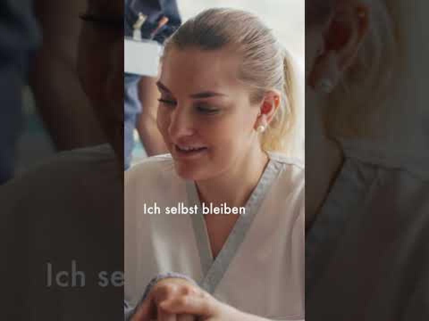 Eine Frau mit nach hinten gebundenen blonden Haaren sitzt in einem Raum und lächelt. Über das Bild ist der deutsche Text „Ich selbst bleiben“ eingeblendet.