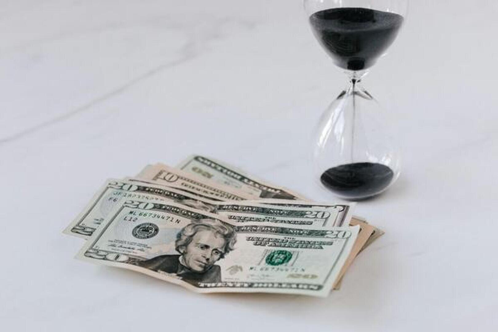 Ein Stapel US-20-Dollar-Scheine liegt auf einer weißen Fläche neben einer Sanduhr mit schwarzem Sand und symbolisiert so das Konzept von Zeit und Geld.