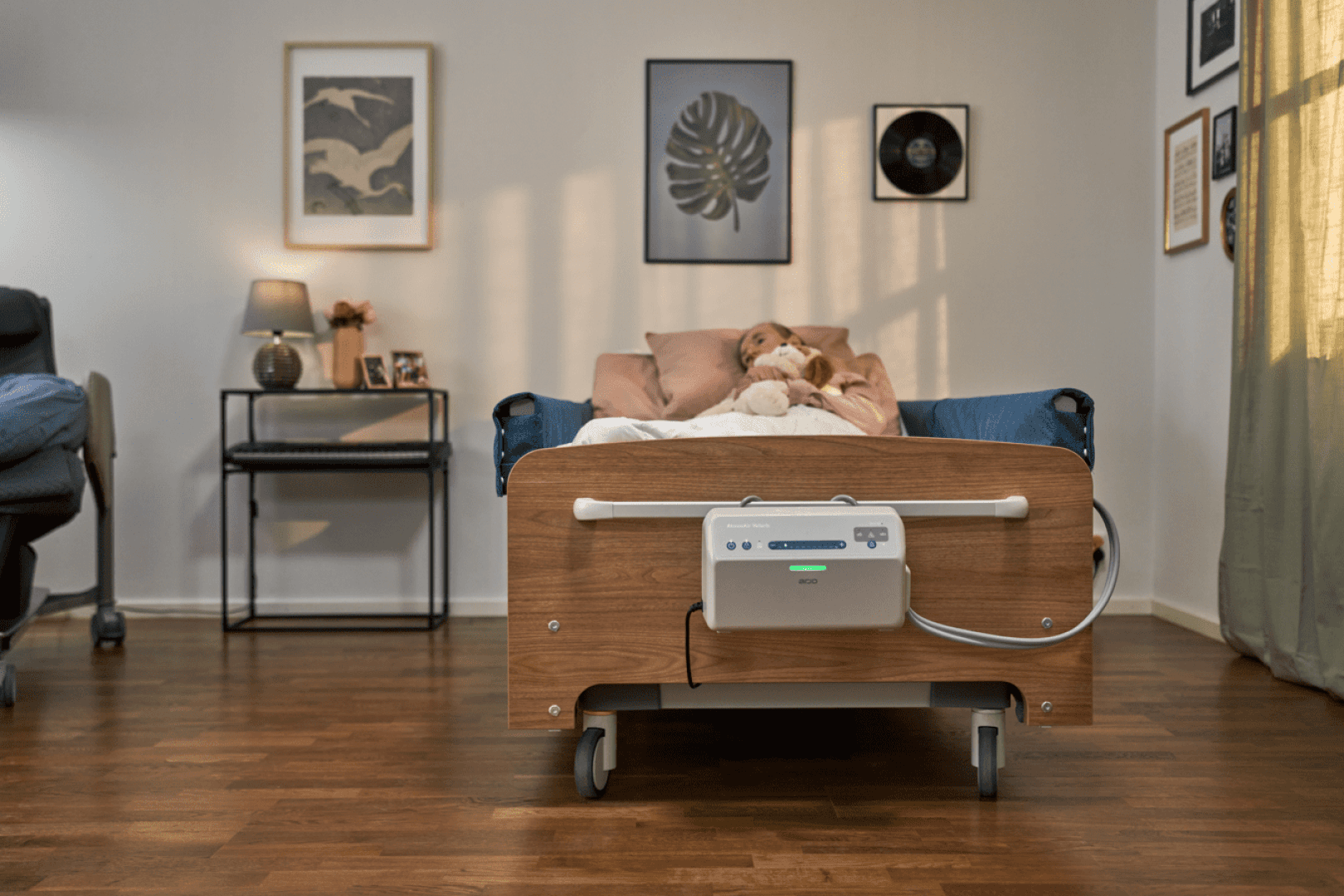 Ein junges Mädchen liegt in einem Krankenhausbett in einem gemütlichen, hell erleuchteten Zimmer mit Holzböden, Wandbildern, einer Lampe auf einem kleinen Tisch und einem Stuhl im Hintergrund.