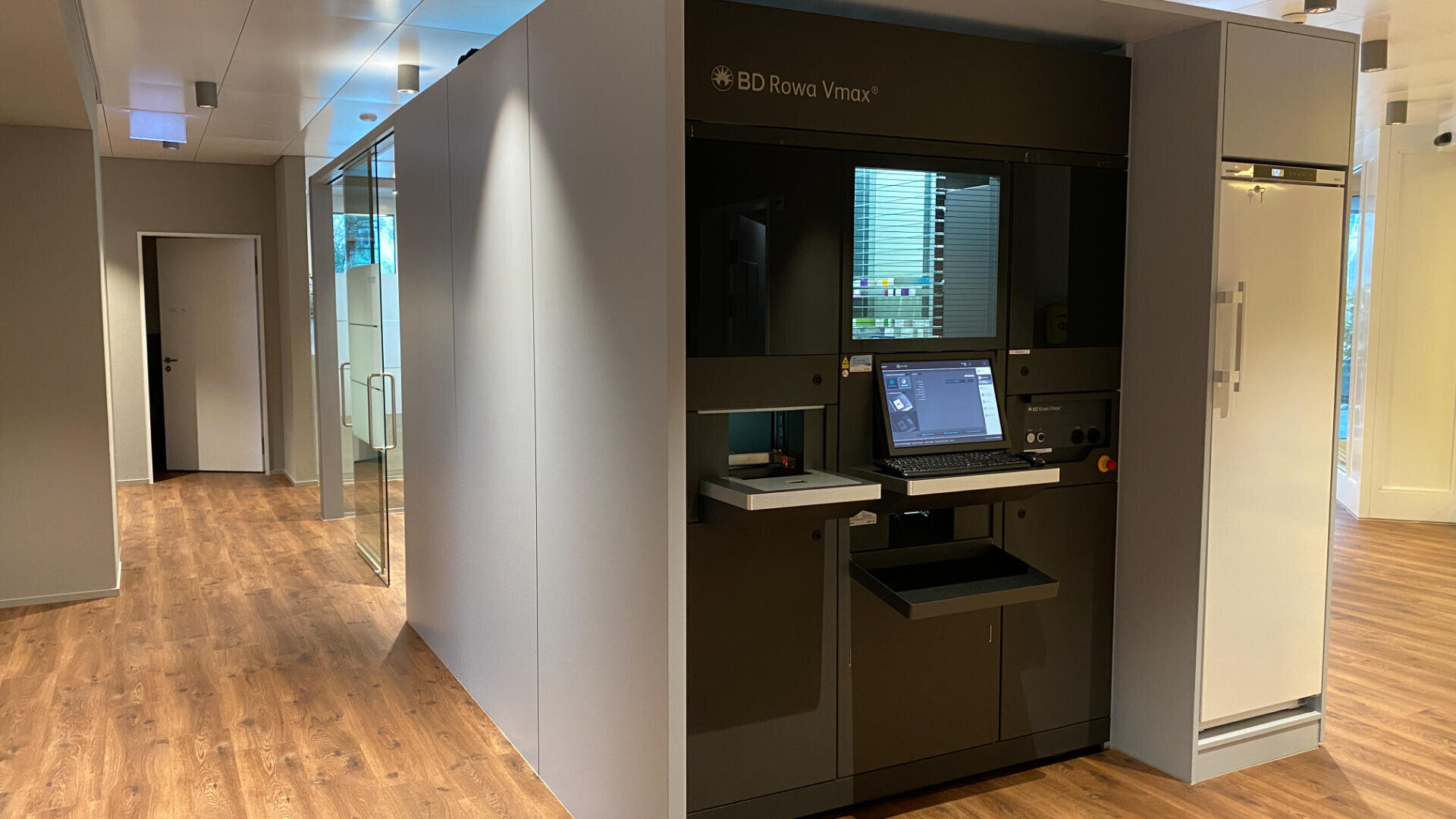 In einem modernen Raum mit Holzfußboden steht ein BD Rowa Vmax, ein automatisches Lager- und Ausgabesystem für Apotheken, neben Büros mit Glaswänden und einem Kühlschrank. Ein Computermonitor und eine Tastatur sind an der Maschine angebracht.