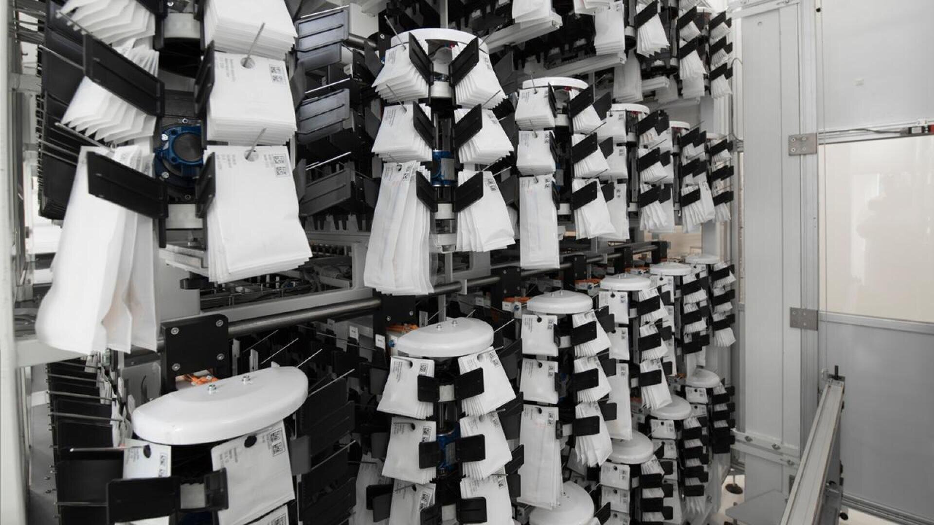 Automatisiertes Sortiersystem mit Reihen von weißen Paketen, die an schwarzen Klammern befestigt sind und auf rotierenden Regalen in einer modernen Industrie- oder Postverarbeitungsanlage angeordnet sind.