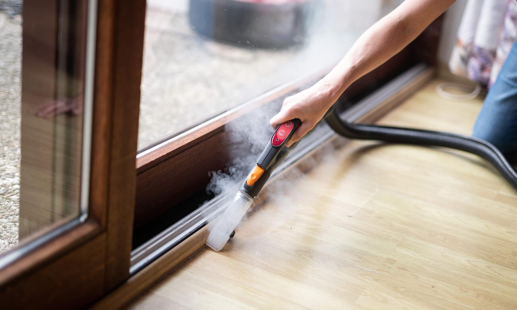 Eine Person verwendet einen Dampfreiniger, um die schmale Schiene einer Glasschiebetür zu reinigen, wobei der Dampf sichtbar aufsteigt, während sie auf einem Holzboden in der Nähe eines Fensters arbeitet.