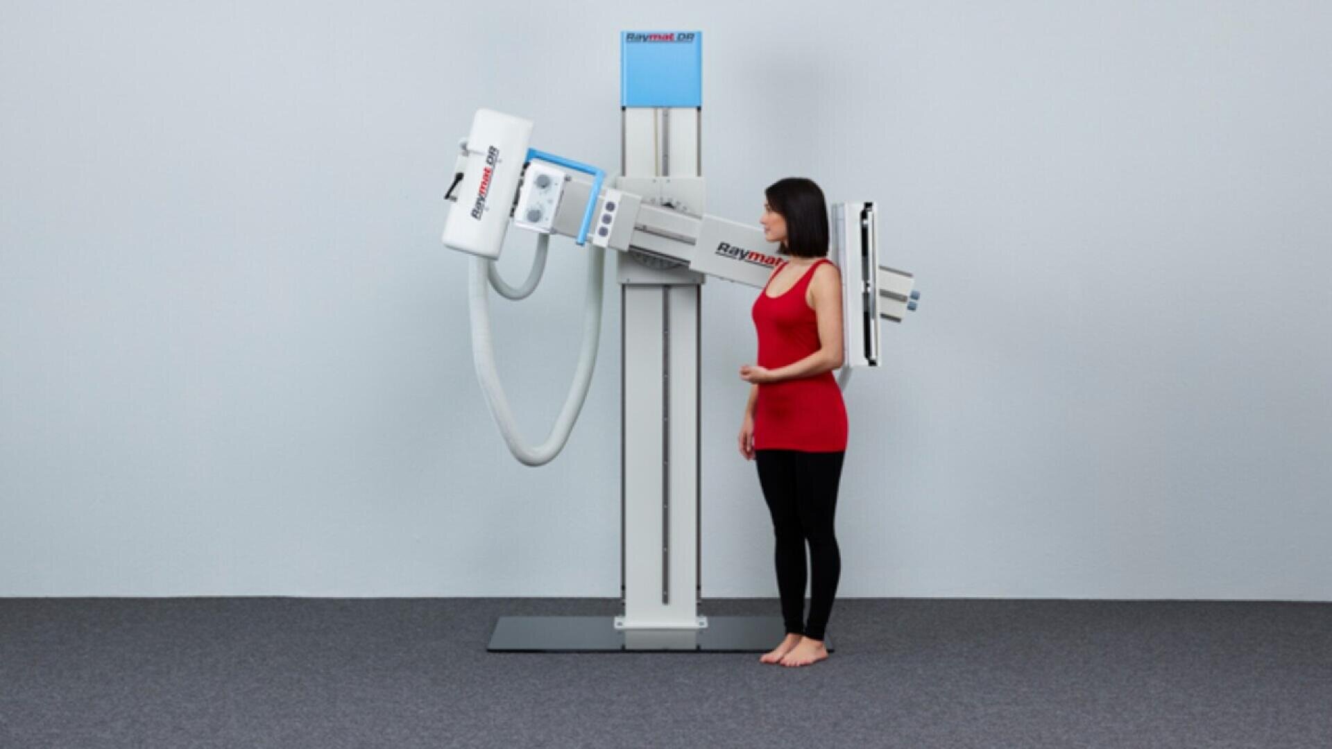 Une femme vêtue d'un haut rouge et d'un legging noir se tient debout à côté d'un appareil de radiographie dans un environnement clinique, positionnée pour un scanner thoracique. La pièce est recouverte d'une moquette grise et les murs sont unis.