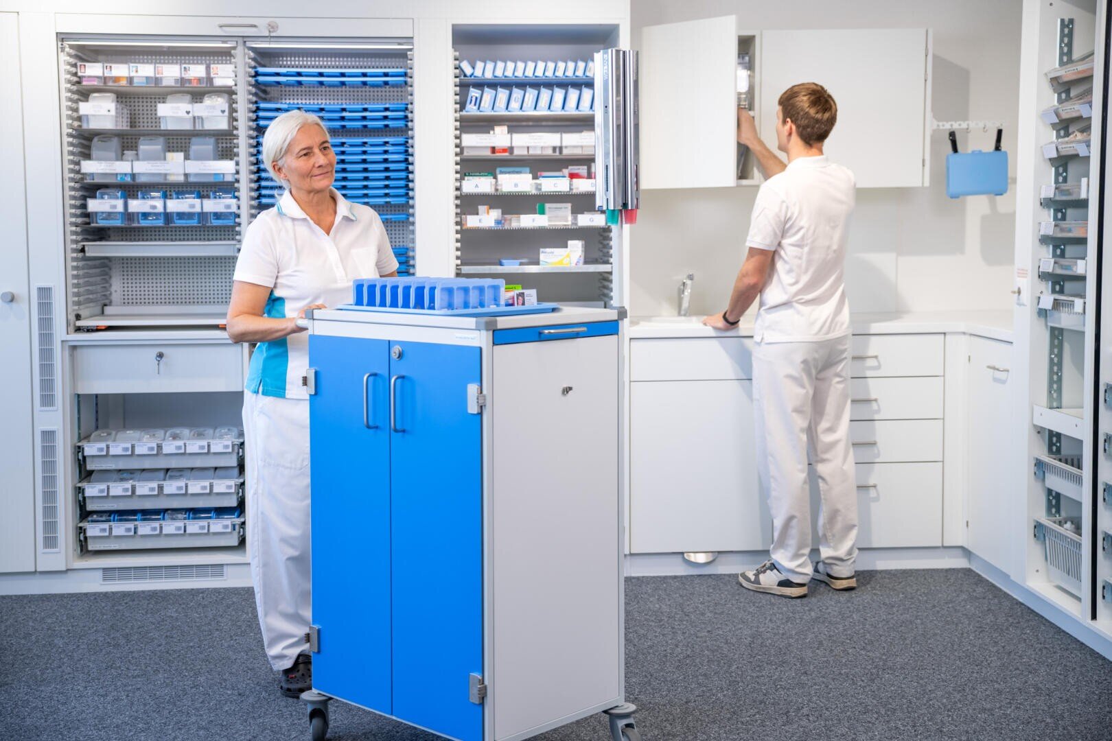 Zwei medizinische Fachkräfte befinden sich in einem Lagerraum für medizinisches Material; die eine steht neben einem Rollwagen, während die andere Gegenstände aus einem Wandschrank entnimmt, der inmitten von Regalen steht, in denen medizinisches Material aufbewahrt wird.