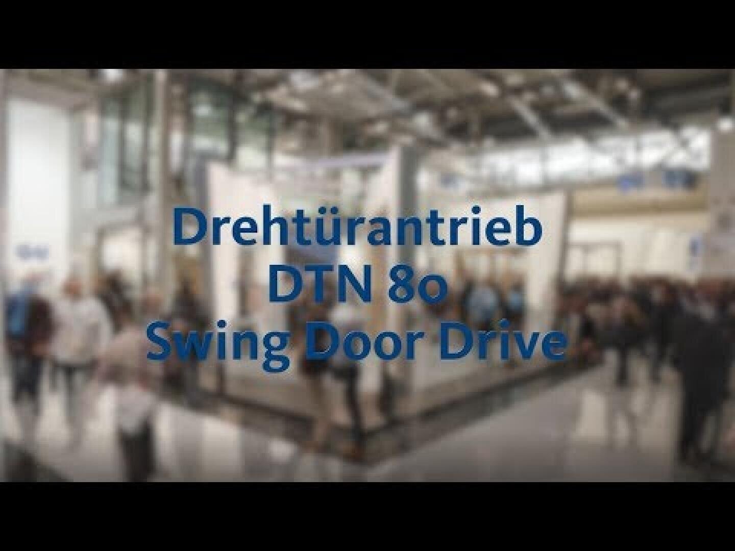 Unscharfes Bild einer Messe oder Ausstellungshalle mit vorbeigehenden Menschen. Der Text in der Mitte lautet: Drehtürantrieb DTN 80 Swing Door Drive.