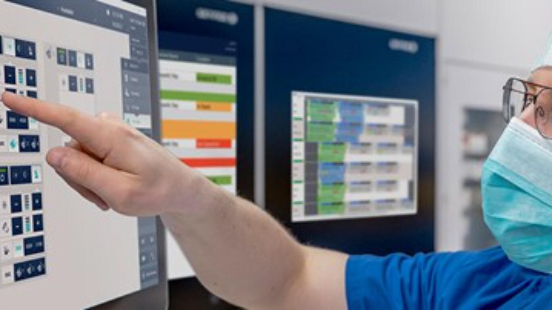 Eine Person, die eine Maske und einen blauen Kittel trägt, zeigt auf einen großen Computerbildschirm, auf dem medizinische oder technische Daten in einem Kontrollraum oder im Gesundheitswesen angezeigt werden.