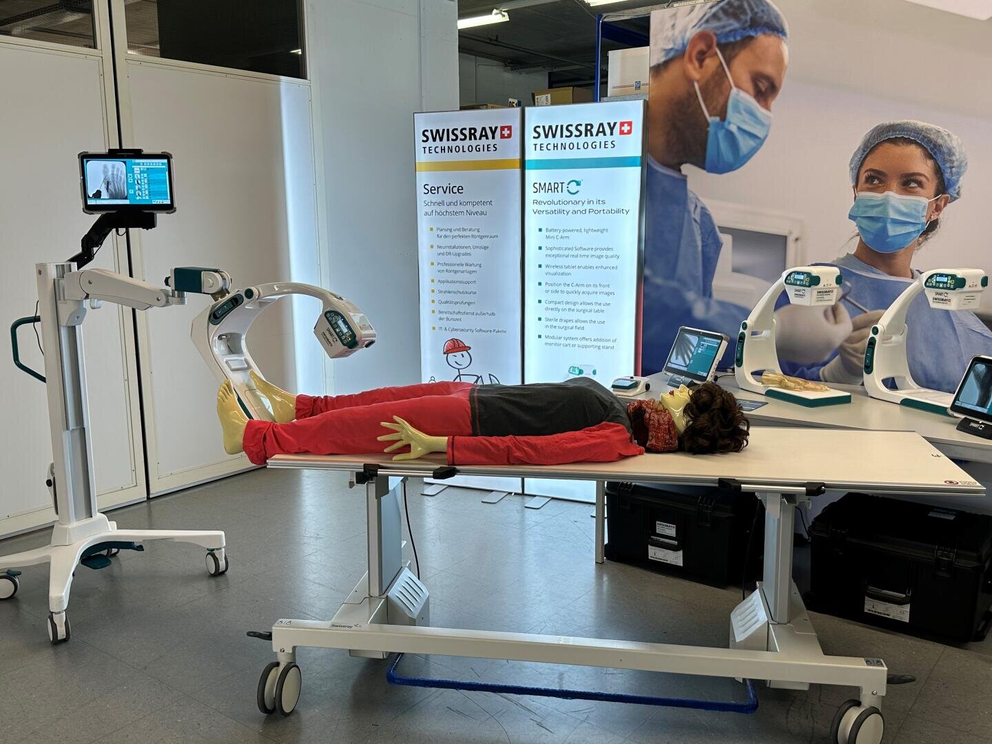 Eine medizinische Übungspuppe liegt auf einem Krankenhausbett mit Beinstützen, umgeben von medizinischen Bildgebungsgeräten. Ein Bildschirm zeigt die Schaufensterpuppe, und im Hintergrund sind Banner von Swissray Technologies zu sehen.