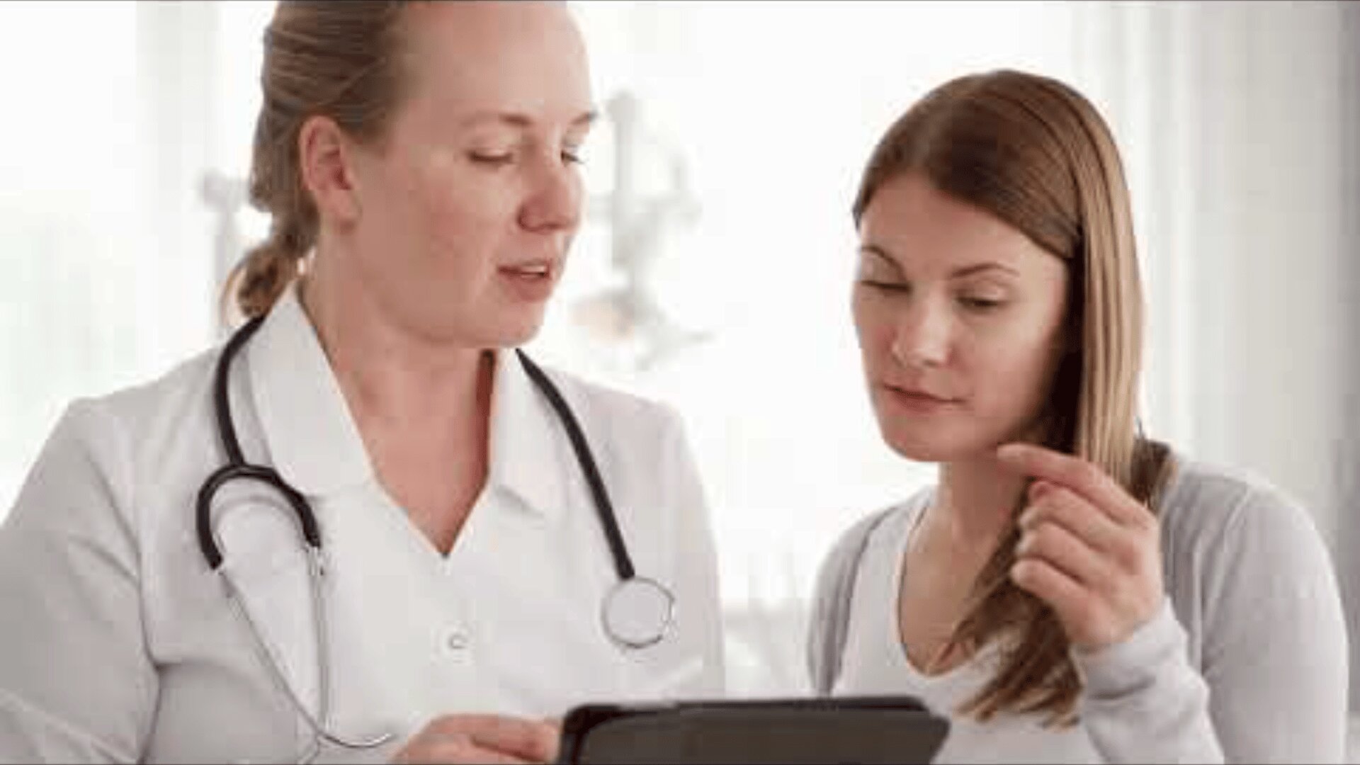Un médecin muni d'un stéthoscope montre une tablette à une femme, lui expliquant des informations dans un cabinet médical très éclairé.