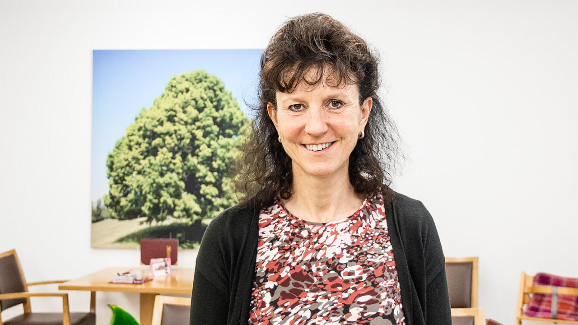 Eine Frau mit lockigem dunklem Haar, die ein gemustertes rot-weißes Oberteil und eine schwarze Strickjacke trägt, steht lächelnd in einem hellen Raum mit Stühlen und einem großen Baumbild an der Wand hinter ihr.