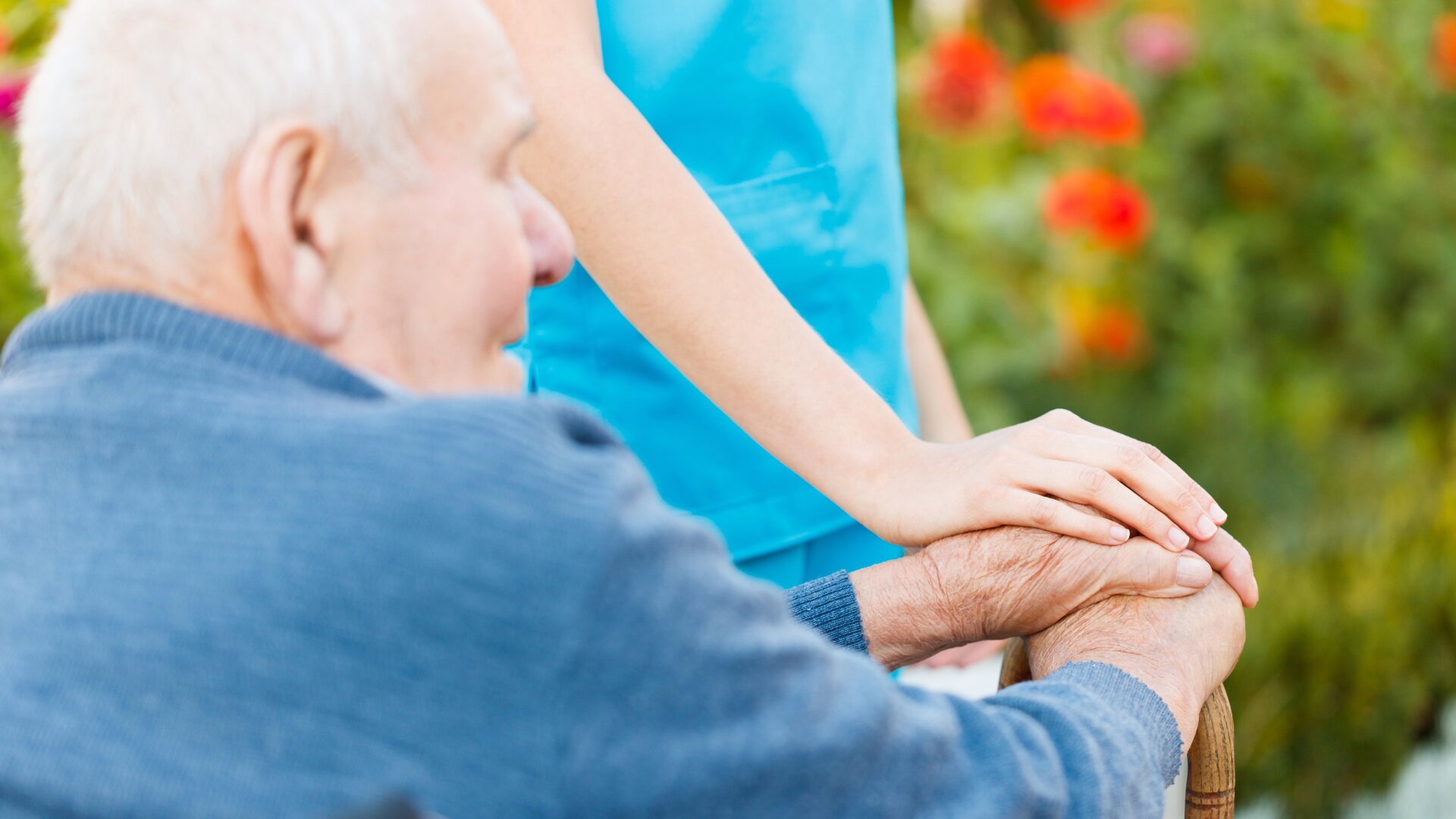 Eine ältere Person sitzt im Freien, während eine Pflegekraft in blauer Kleidung ihre Hand sanft auf die Hände der älteren Person legt, um sie zu trösten und zu unterstützen. Helle, verschwommene Blumen erscheinen im Hintergrund.