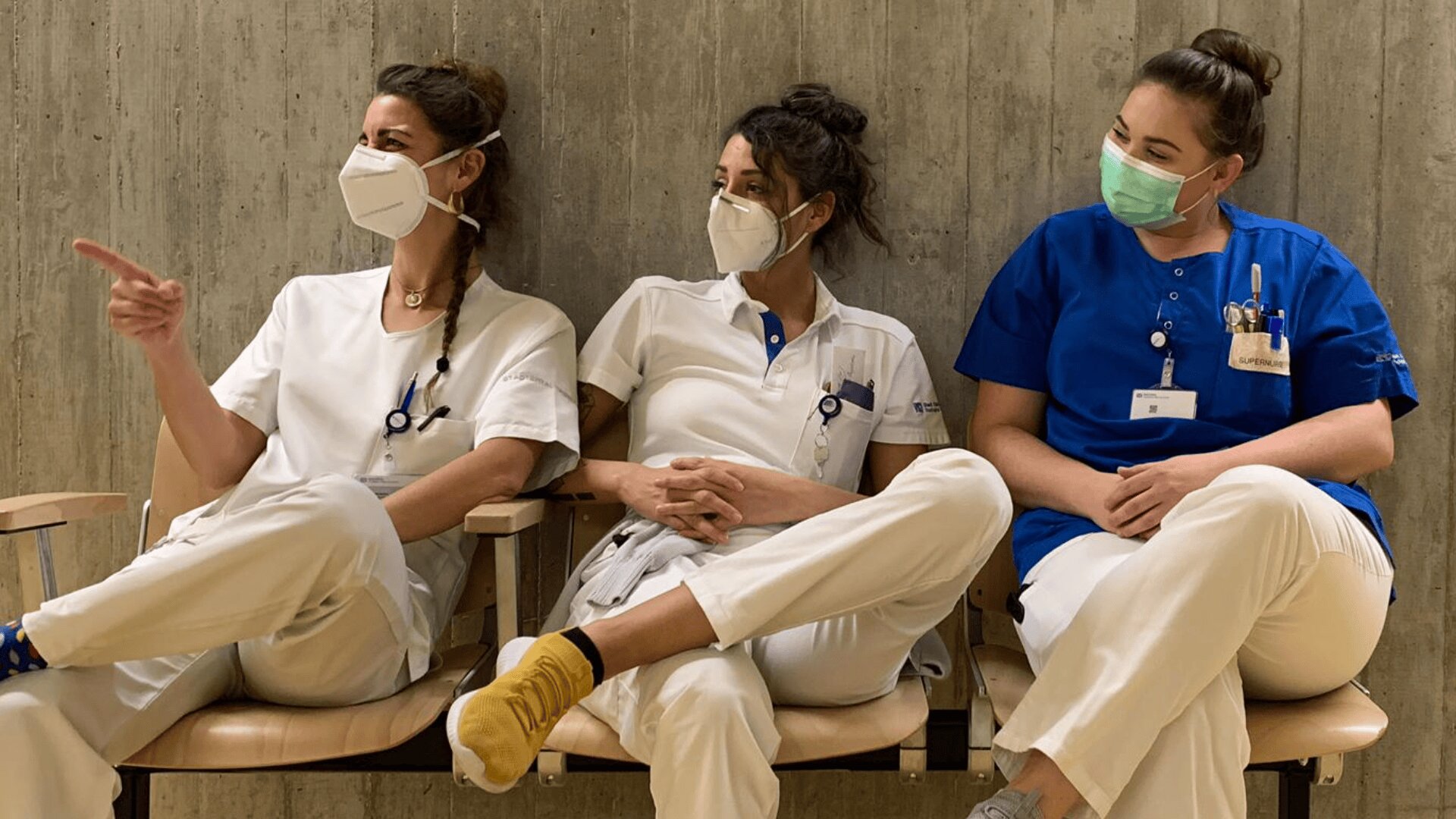 Drei Mitarbeiter des Gesundheitswesens mit Masken, zwei in weißen und einer in blauen Uniformen, sitzen auf Stühlen an einer Betonwand. Sie wirken entspannt, einer zeigt auf sie und die anderen schauen in dieselbe Richtung.