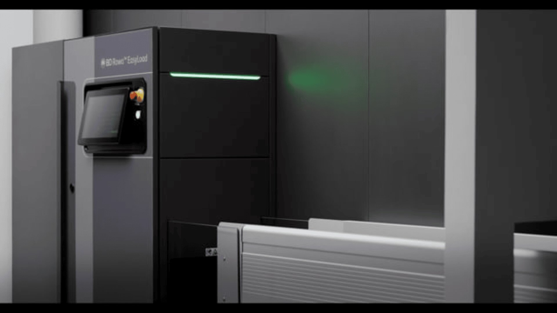 Eine schlanke, moderne Industriemaschine mit einem Touchscreen-Display und Bedientasten, die vor einer dunklen Wand steht und vor der ein horizontales Metalltablett oder ein Förderband steht.