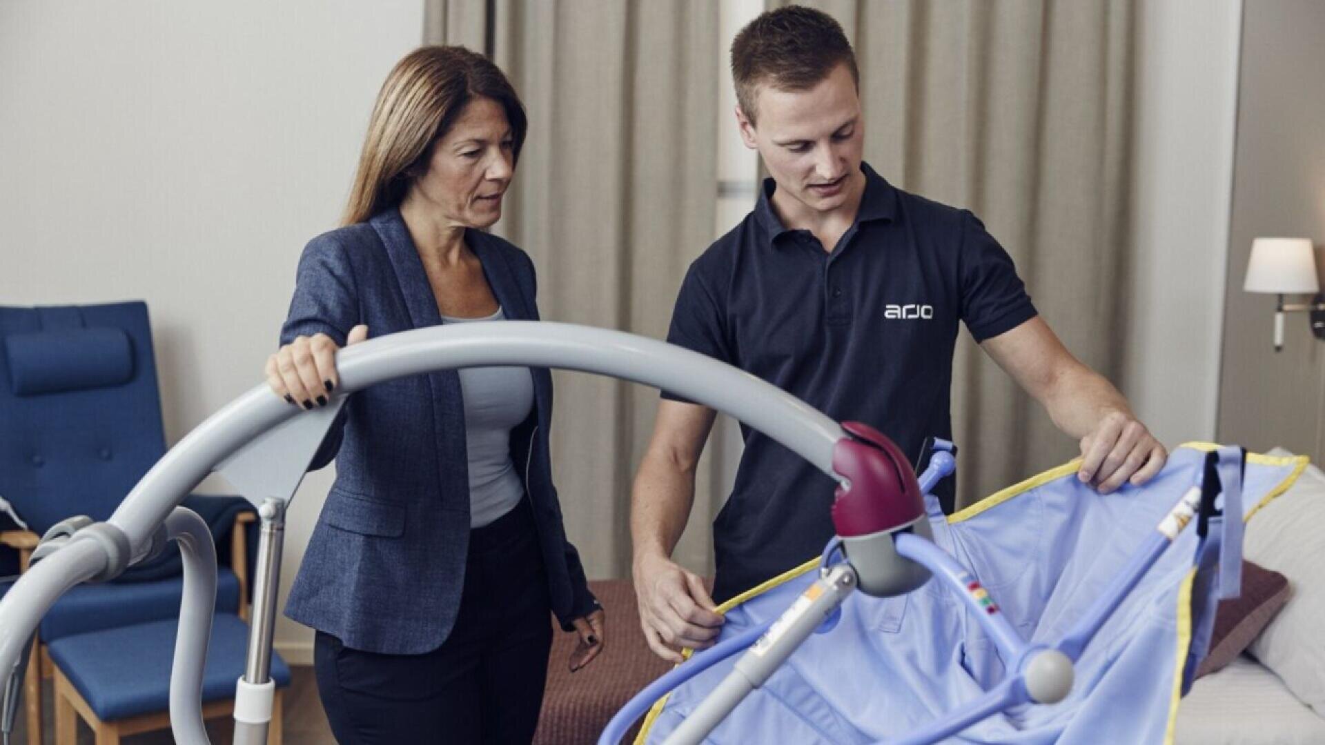 Une femme et un homme se tiennent à côté d'un lit d'hôpital et examinent un équipement médical avec un harnais bleu, peut-être un lève-personne. L'homme explique ses caractéristiques tandis que la femme écoute attentivement.