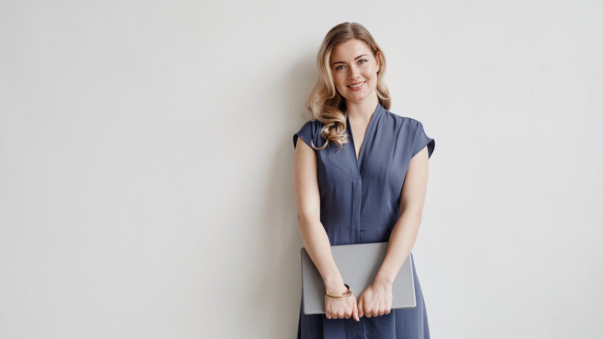 Eine Frau mit langen blonden Haaren und einem blauen Kleid steht vor einer schlichten weißen Wand, lächelt und hält einen geschlossenen Laptop mit beiden Händen vor sich.