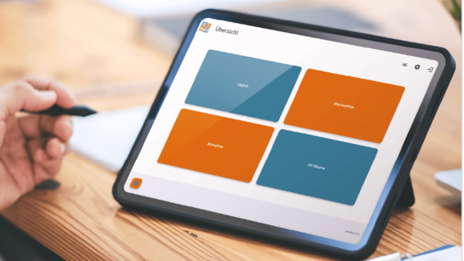 Eine Person hält einen Stift in der Nähe eines Tablets, auf dem ein Armaturenbrett mit vier farbigen, in deutscher Sprache beschrifteten Feldern auf einem Holztisch steht.