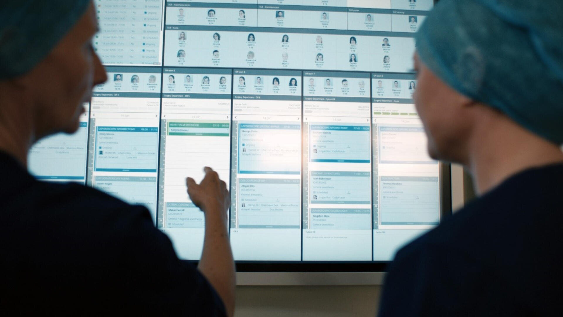 Zwei Personen in Kitteln und Mützen prüfen und besprechen Informationen, die auf einem großen digitalen Bildschirm mit medizinischen oder terminlichen Daten angezeigt werden.
