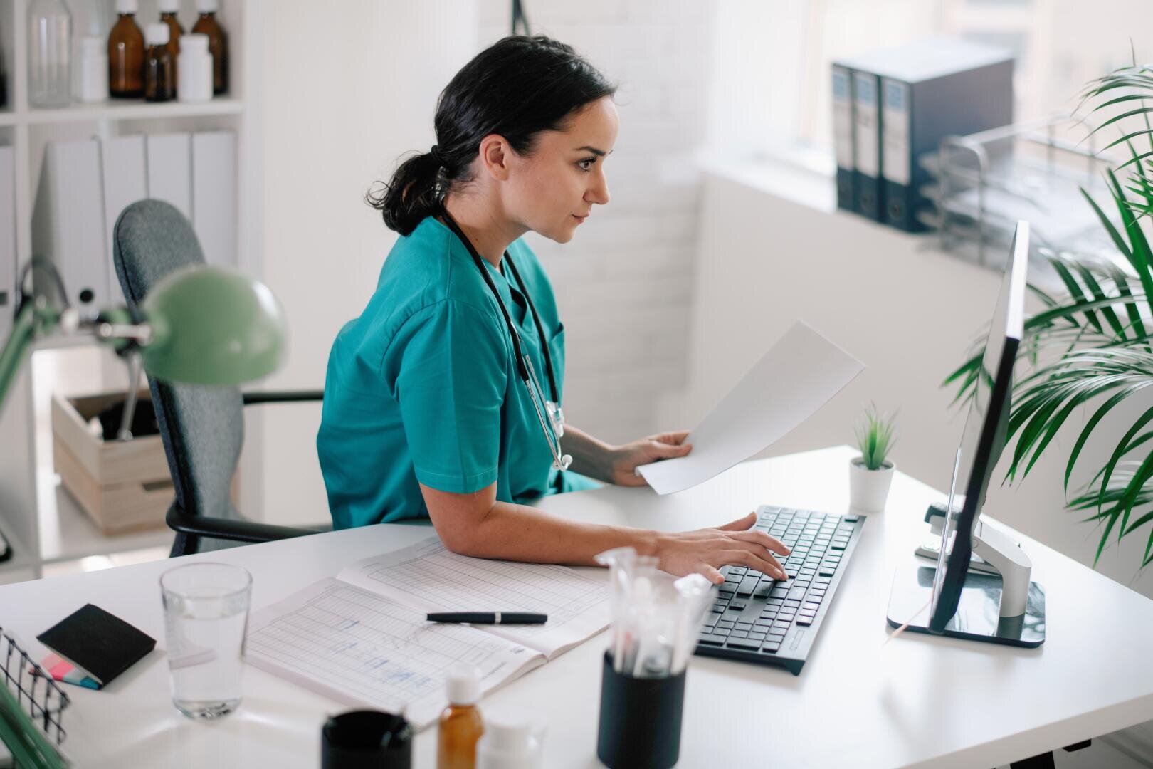 Ein Mitarbeiter im Gesundheitswesen in OP-Kleidung sitzt an einem Schreibtisch, hält Unterlagen in der Hand und tippt auf einer Computertastatur in einem hellen Büro, in dem Krankenakten, Pflanzen und Arbeitsutensilien zu sehen sind.