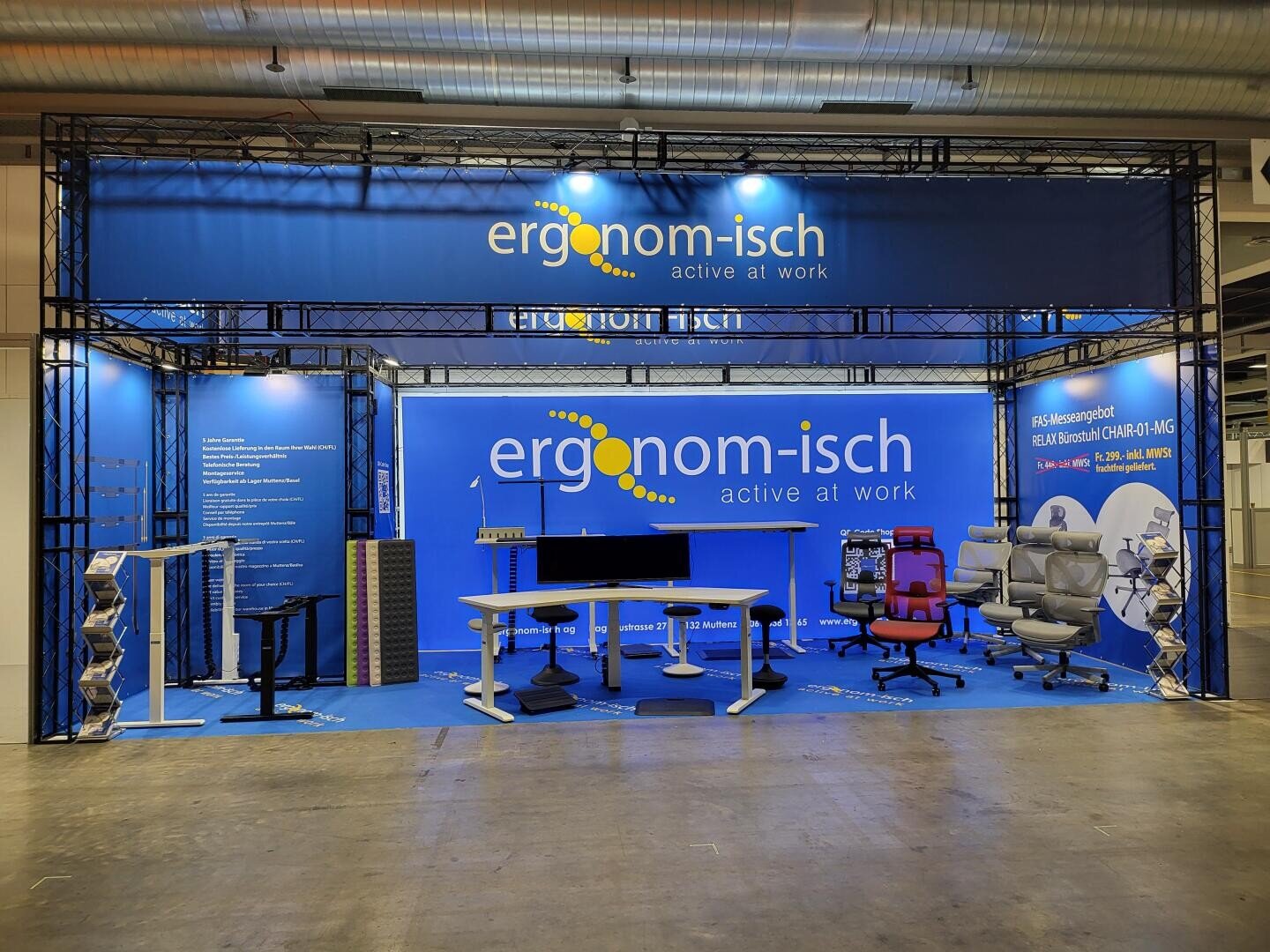 Messestand für „ergonom-isch“ mit ergonomischen Büromöbeln, darunter Stühle, Schreibtische und Zubehör, mit blauem Branding und Informationsbannern. Der Stand ist gut beleuchtet und für Besucher übersichtlich gestaltet.