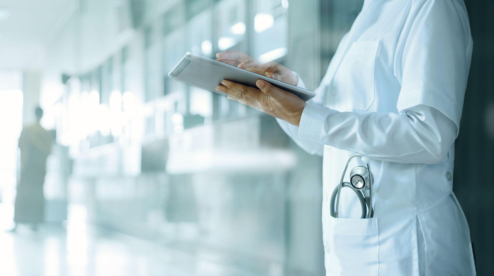 Ein Arzt in einem weißen Kittel steht mit einem Tablet in der Hand in einem Krankenhausflur, ein Stethoskop steckt in seiner Tasche, und im Hintergrund ist eine weitere Person unscharf zu erkennen.