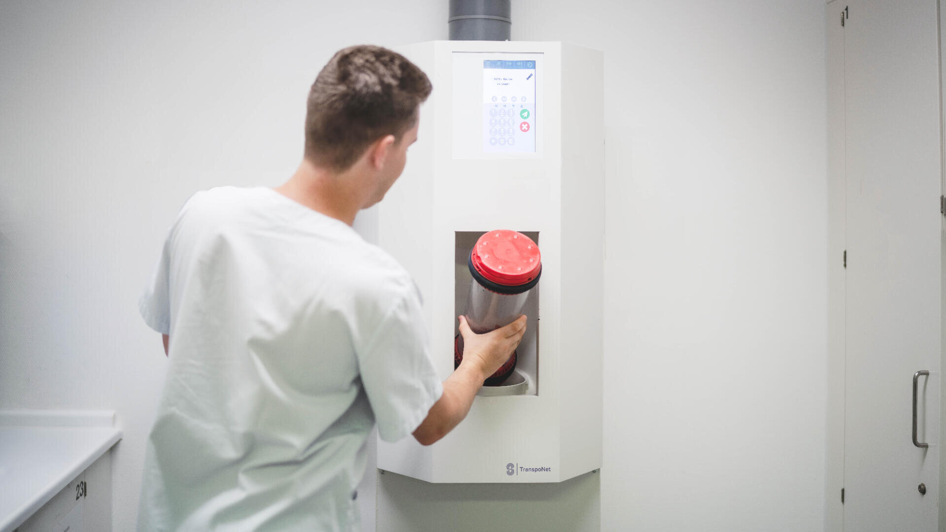 Eine Person in medizinischer Kleidung setzt einen zylindrischen Behälter mit rotem Deckel in ein medizinisches Gerät ein, das in einer klinischen Umgebung an der Wand befestigt ist.