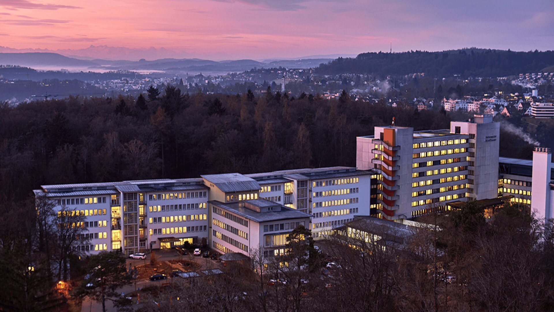 Ein großer Krankenhauskomplex mit beleuchteten Fenstern, umgeben von Bäumen, liegt unter einem violett-rosa Sonnenuntergangshimmel, im Hintergrund sind die Berge und eine Stadt zu sehen.