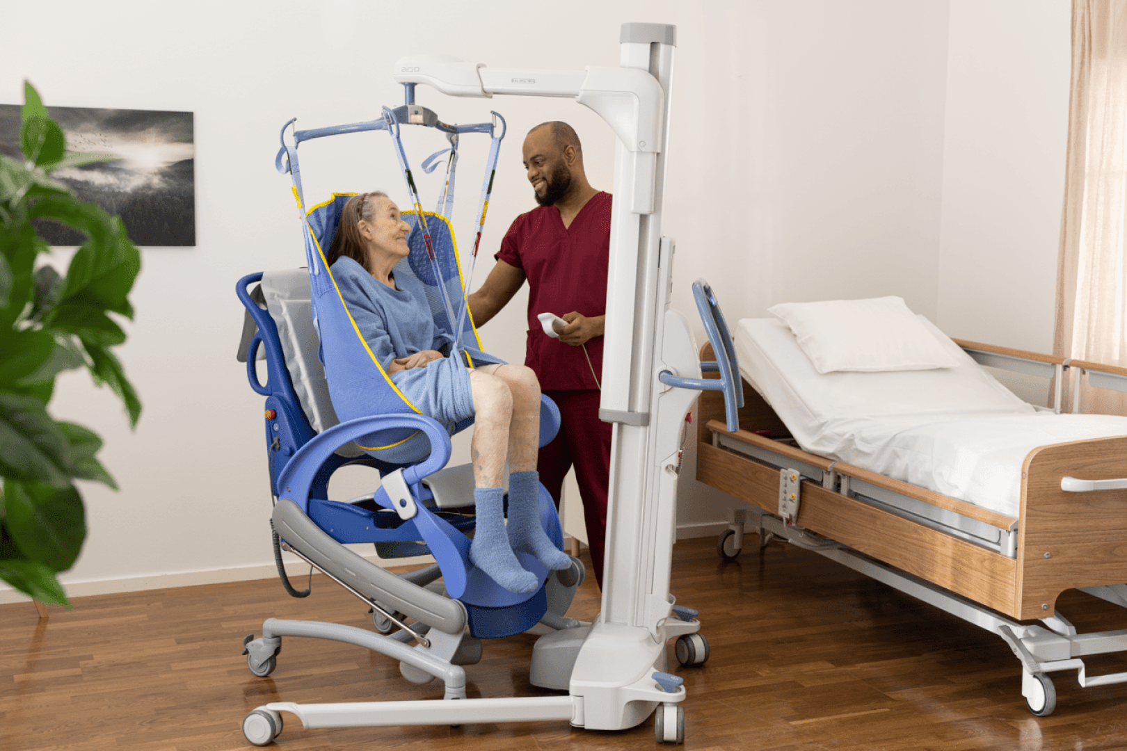 Une aide-soignante en tenue chirurgicale aide une femme âgée, assise dans un fauteuil élévateur, dans une pièce très éclairée où se trouvent un lit d'hôpital et une plante.