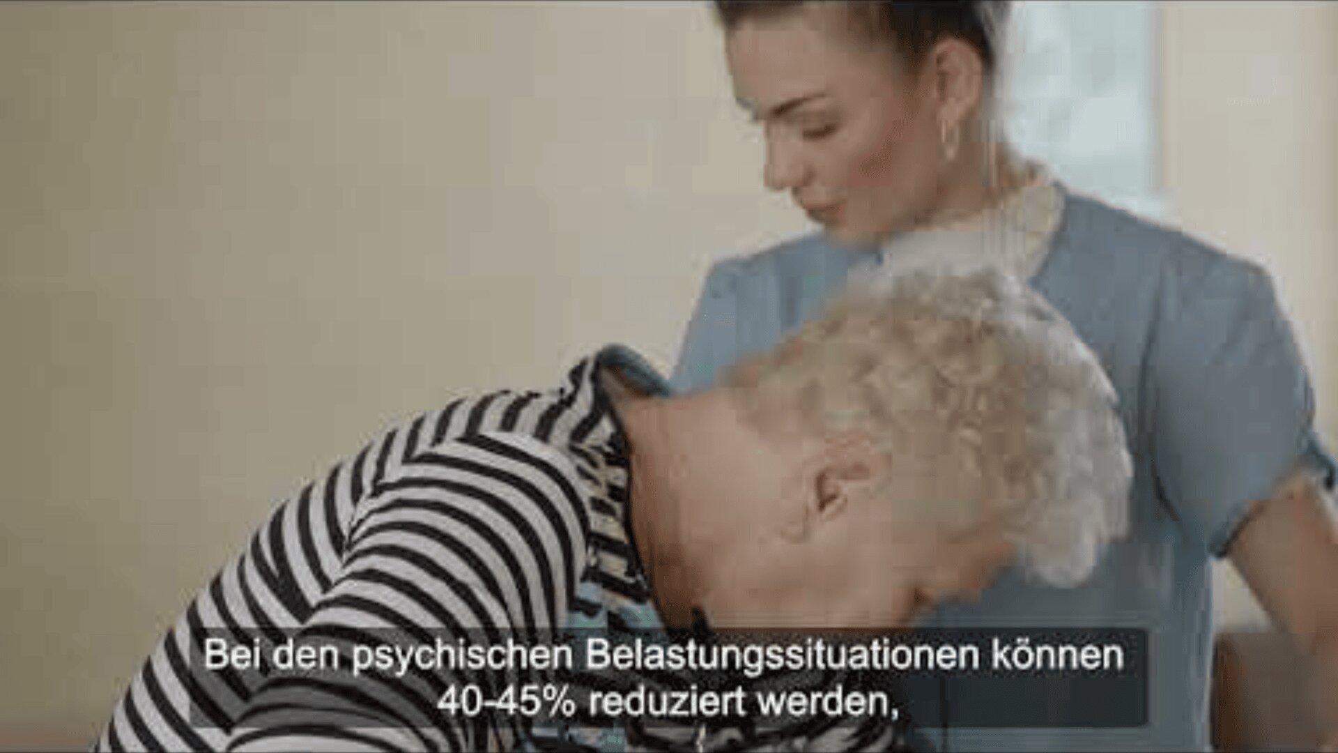 Une infirmière en blouse bleue soutient une personne âgée aux cheveux blancs portant une chemise à rayures noires et blanches. Des sous-titres en allemand sont visibles en bas de l'image.