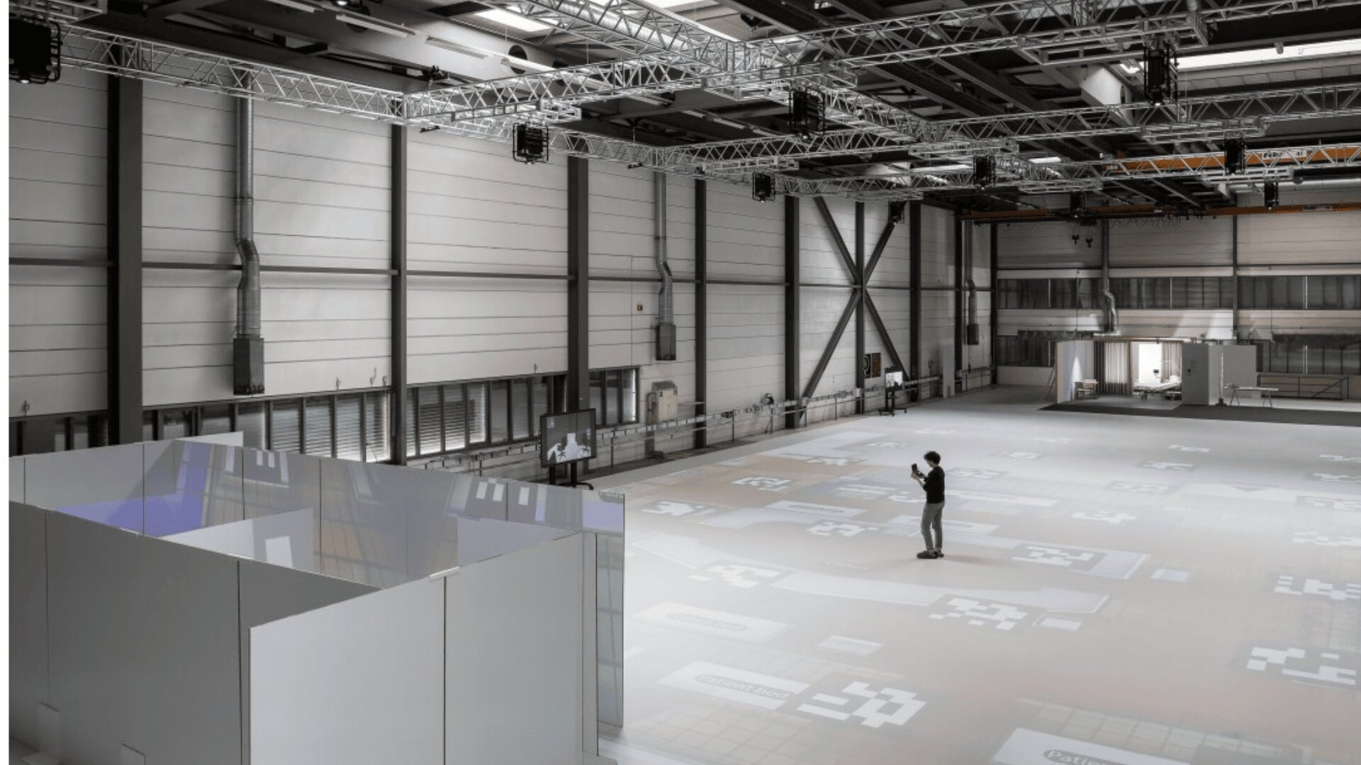 Une personne se tient seule dans un grand espace industriel intérieur avec de hauts plafonds et des poutres métalliques, entourée de projections numériques sur le sol et les murs, avec des écrans et des zones cloisonnées visibles.