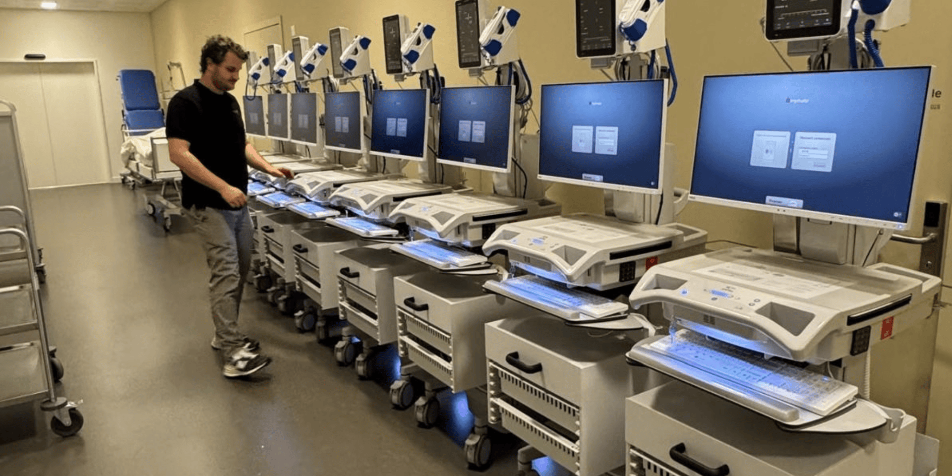 Eine Person steht neben einer Reihe von medizinischen Computerarbeitsplätzen mit großen Monitoren und Tastaturen in einer medizinischen Einrichtung, möglicherweise einem Krankenhaus oder einer Klinik.