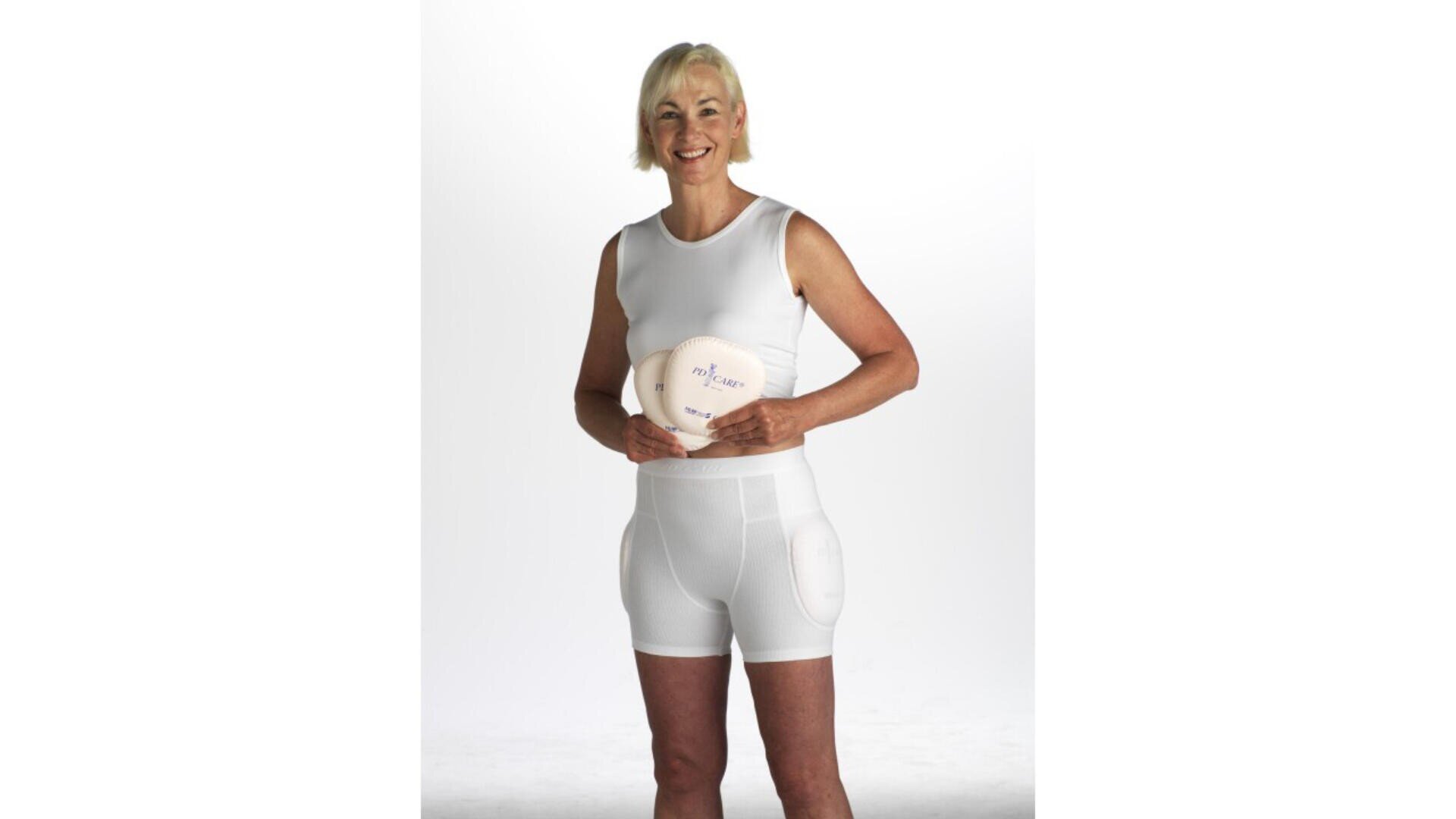 Une femme âgée aux cheveux blonds courts sourit et pose dans un haut blanc sans manches et un short rembourré, tenant un ballon de volley-ball blanc sur un fond uni et lumineux.