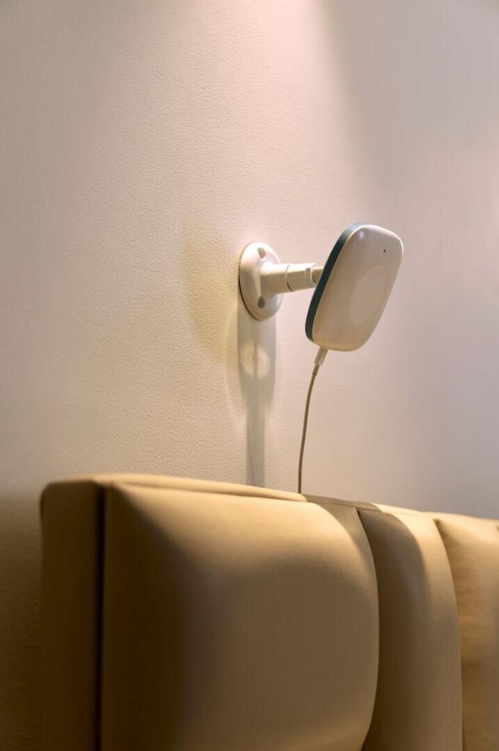 Un petit capteur intelligent blanc et rectangulaire fixé au mur au-dessus d'une tête de lit rembourrée, avec un câble d'alimentation qui pend.