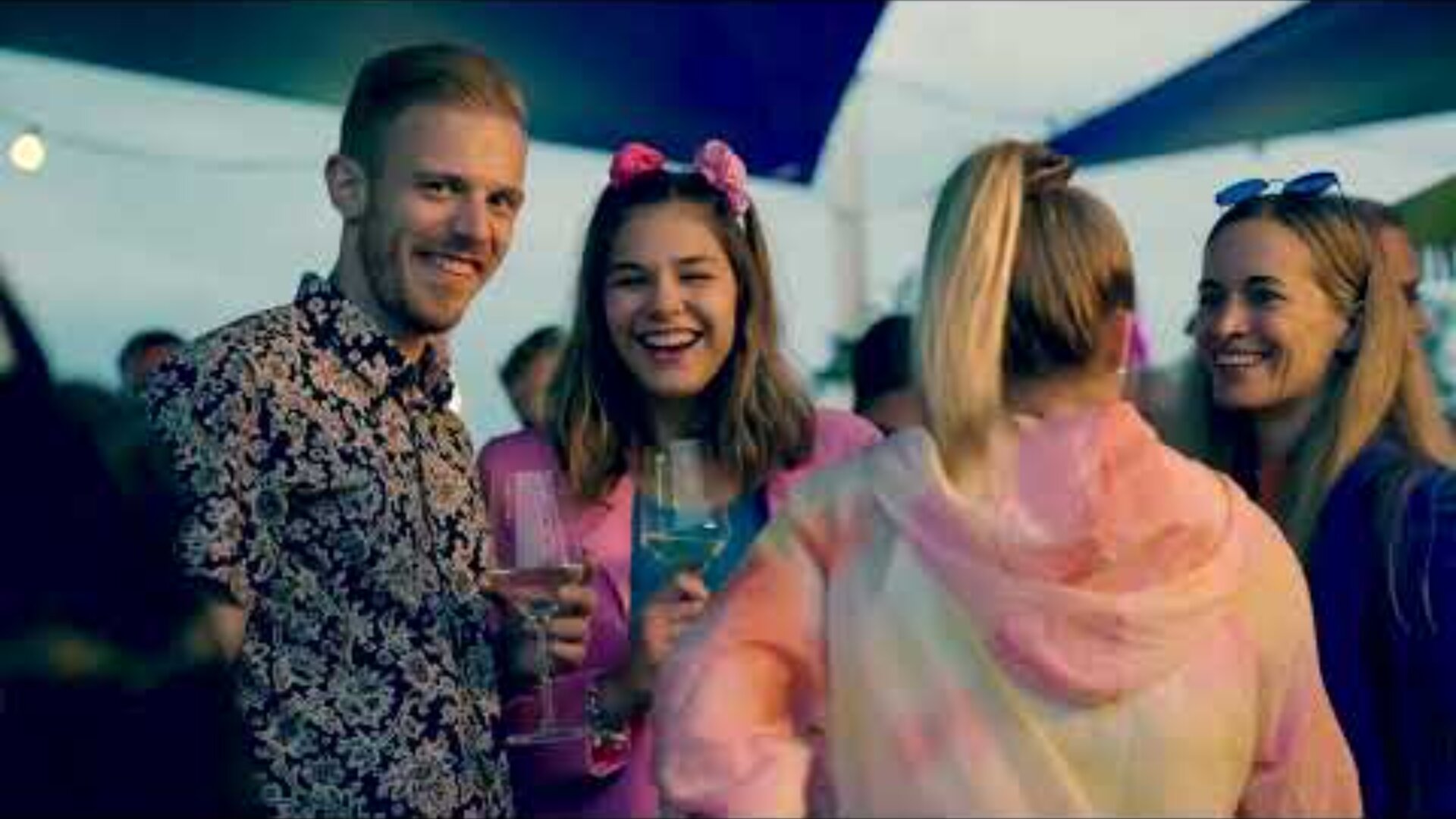 Quatre jeunes adultes se tiennent côte à côte lors d'un événement en plein air, souriant et tenant des boissons. Les vêtements et les décorations colorés suggèrent une atmosphère festive et joyeuse.