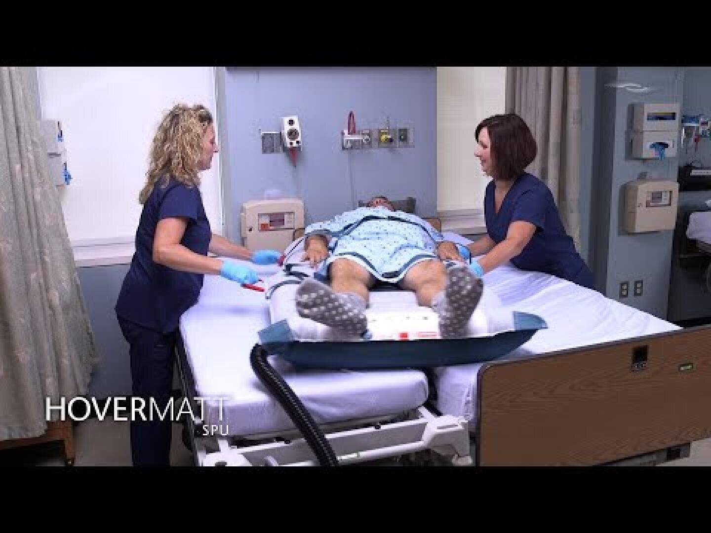 Deux professionnels de santé en tenue de bloc utilisent un système de transfert pneumatique HoverMatt pour déplacer un patient couché sur un lit d'hôpital dans une salle de soins. Le logo « HOVERMATT » est visible dans le coin inférieur gauche.