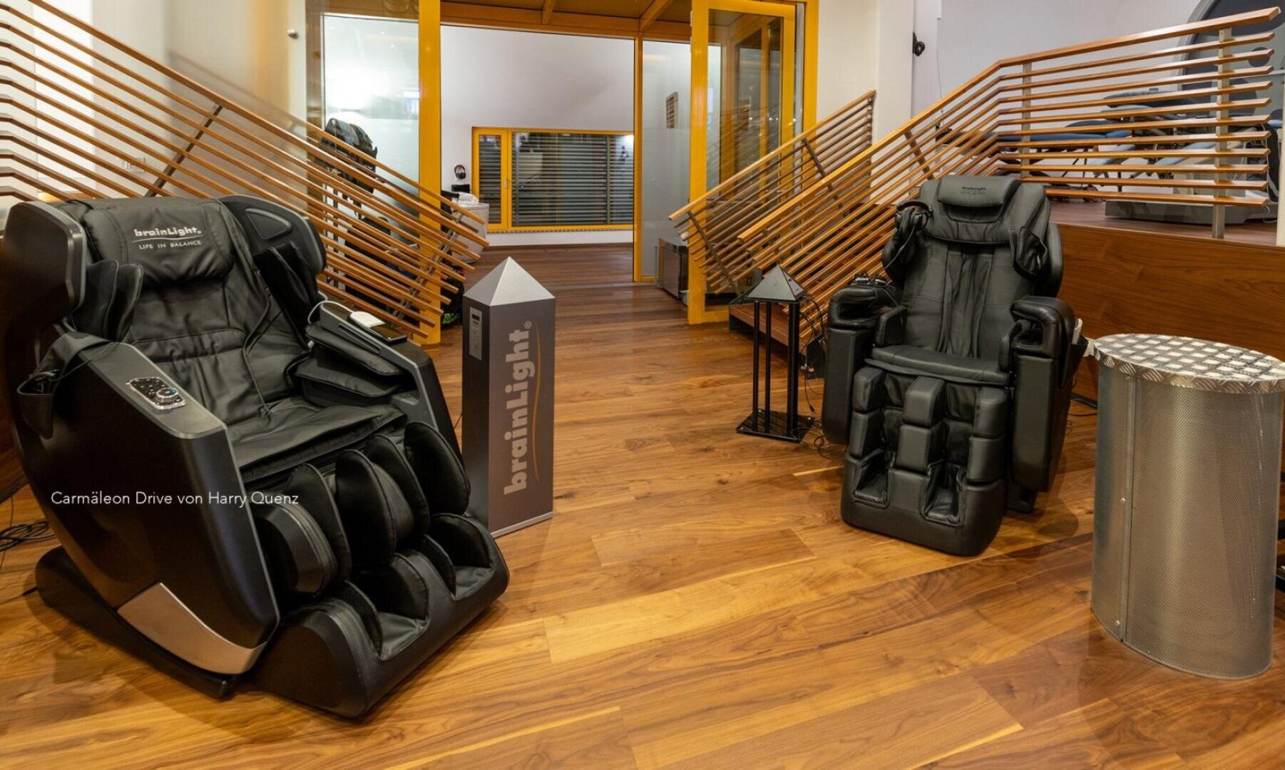 Zwei schwarze Massagesessel der Marke brainLight stehen auf einem Parkettboden in einer Eingangshalle, in der sich eine dekorative Treppe mit Holzlamellen befindet.