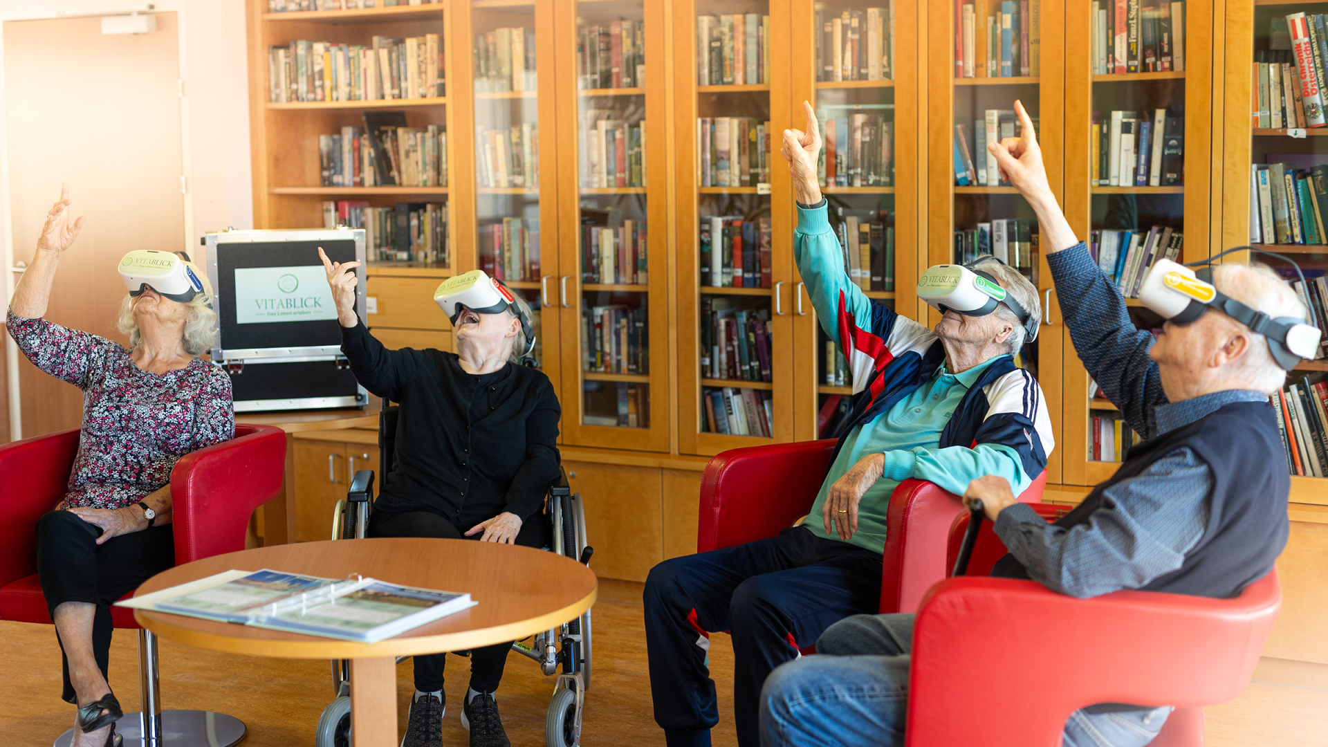 Vier Senioren, die Vitablick-VR-Headsets tragen, sitzen in einer Bibliothek und strecken jeweils einen Arm zur Decke.