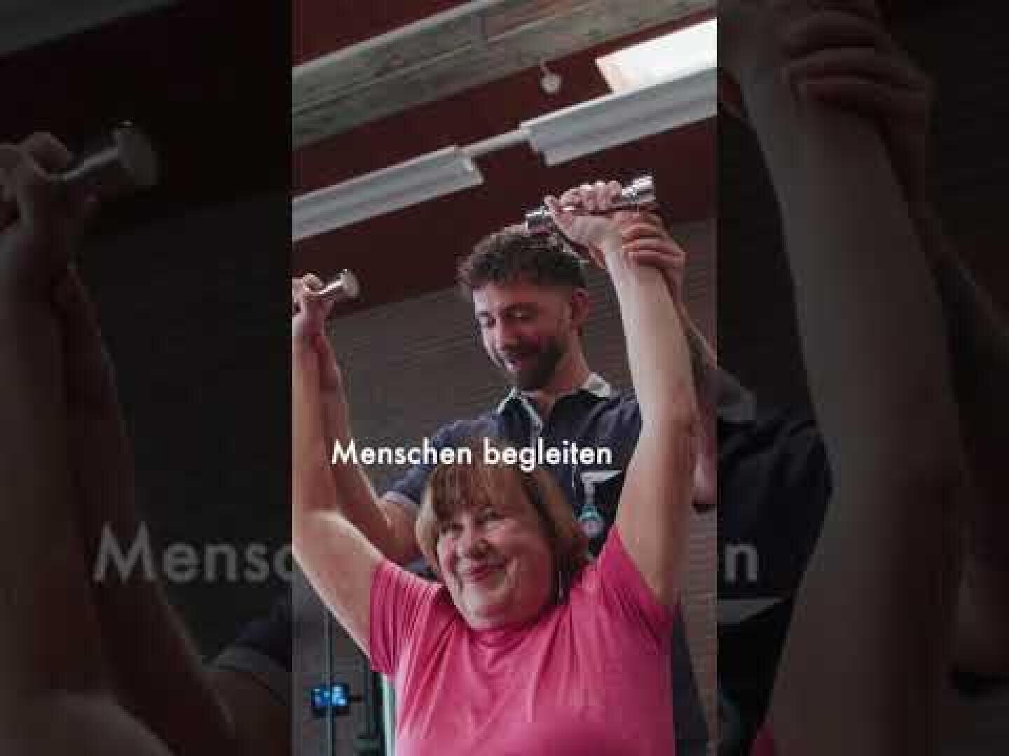 Eine Frau lächelt und hebt die Arme, während ein Mann hinter ihr steht; beide halten kleine Hanteln in den Händen. Der deutsche Text lautet: „Menschen begleiten“. Der Schauplatz scheint ein Fitnessstudio oder eine Physiotherapiepraxis zu sein.
