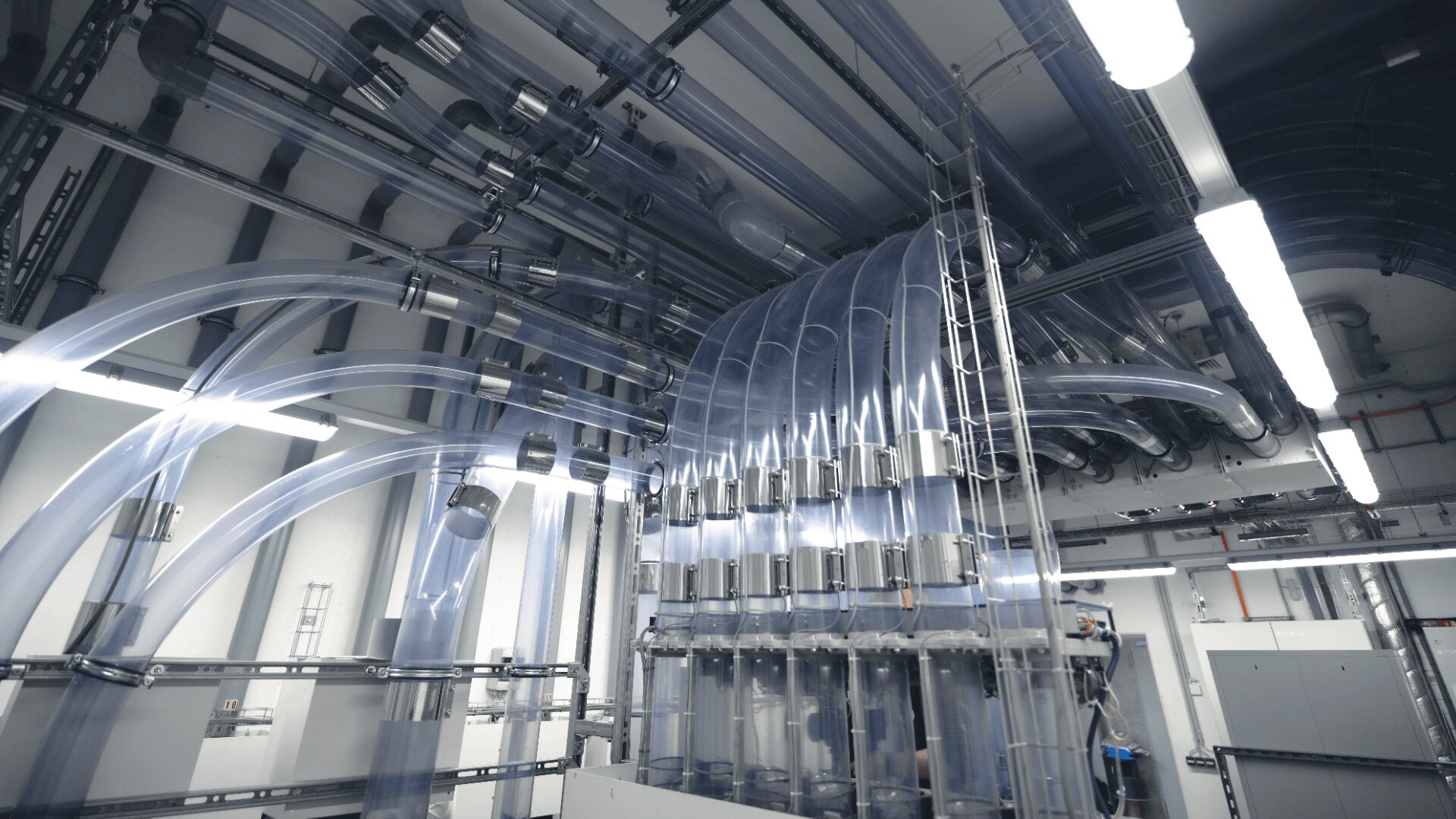 An der Decke und den Wänden eines hell erleuchteten Industrieraums verläuft ein Netz großer transparenter Röhren, die mit verschiedenen Maschinen und Geräten darunter verbunden sind.