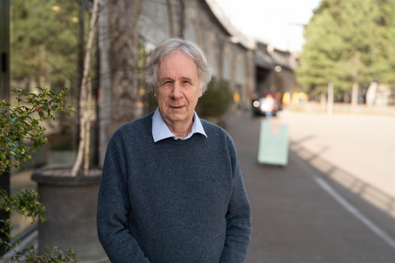 Ein älterer Mann mit grauen Haaren, der einen blauen Pullover und ein Hemd mit Kragen trägt, steht an einem sonnigen Tag draußen in der Nähe eines Gebäudes und einiger Bäume. Im Hintergrund sind die Straße und ein unscharfes Schild zu sehen.