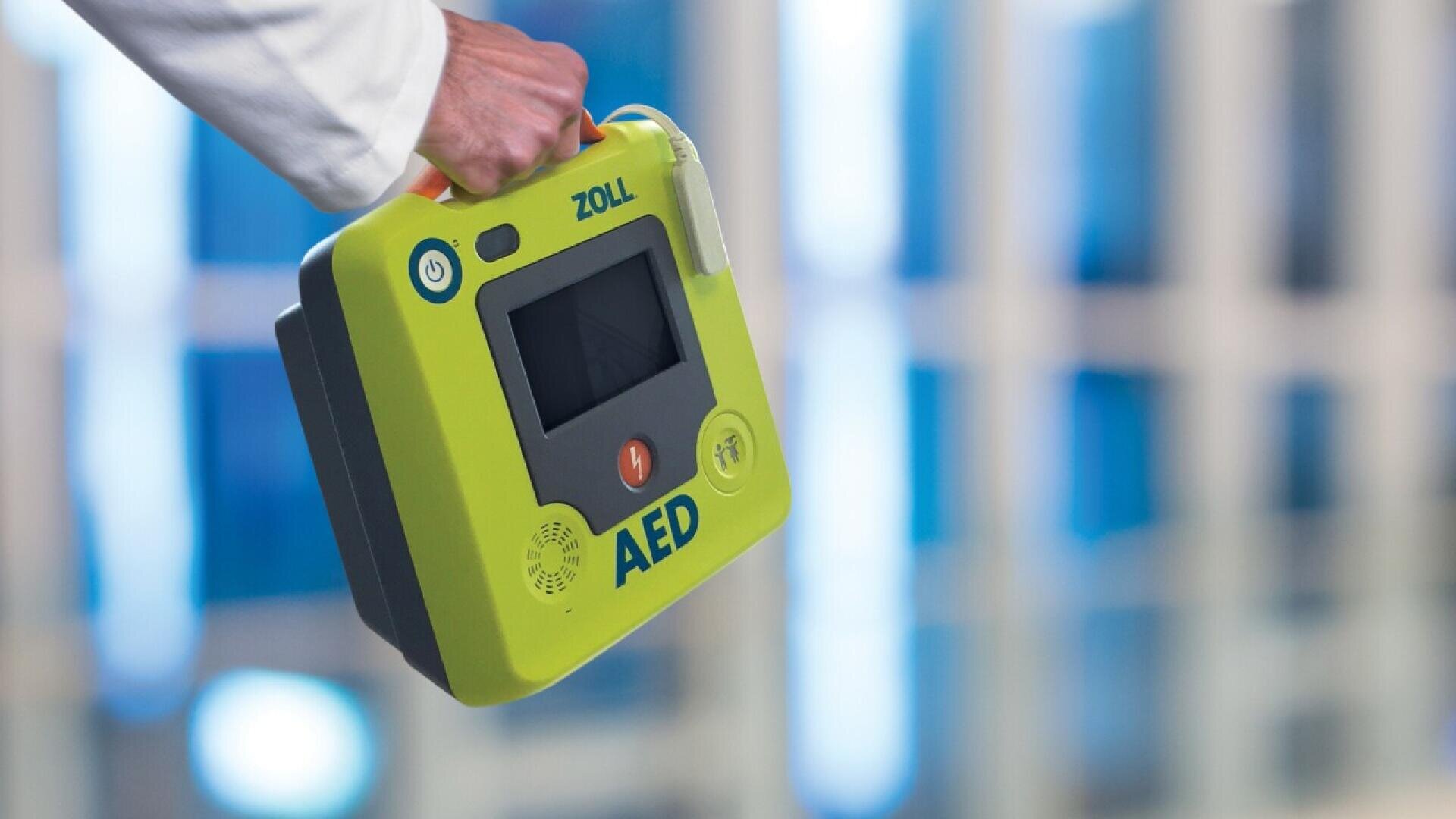 Une personne tenant un défibrillateur externe automatisé (DEA) portable de marque ZOLL par sa poignée jaune, sur un fond flou de fenêtres ou de panneaux de verre.