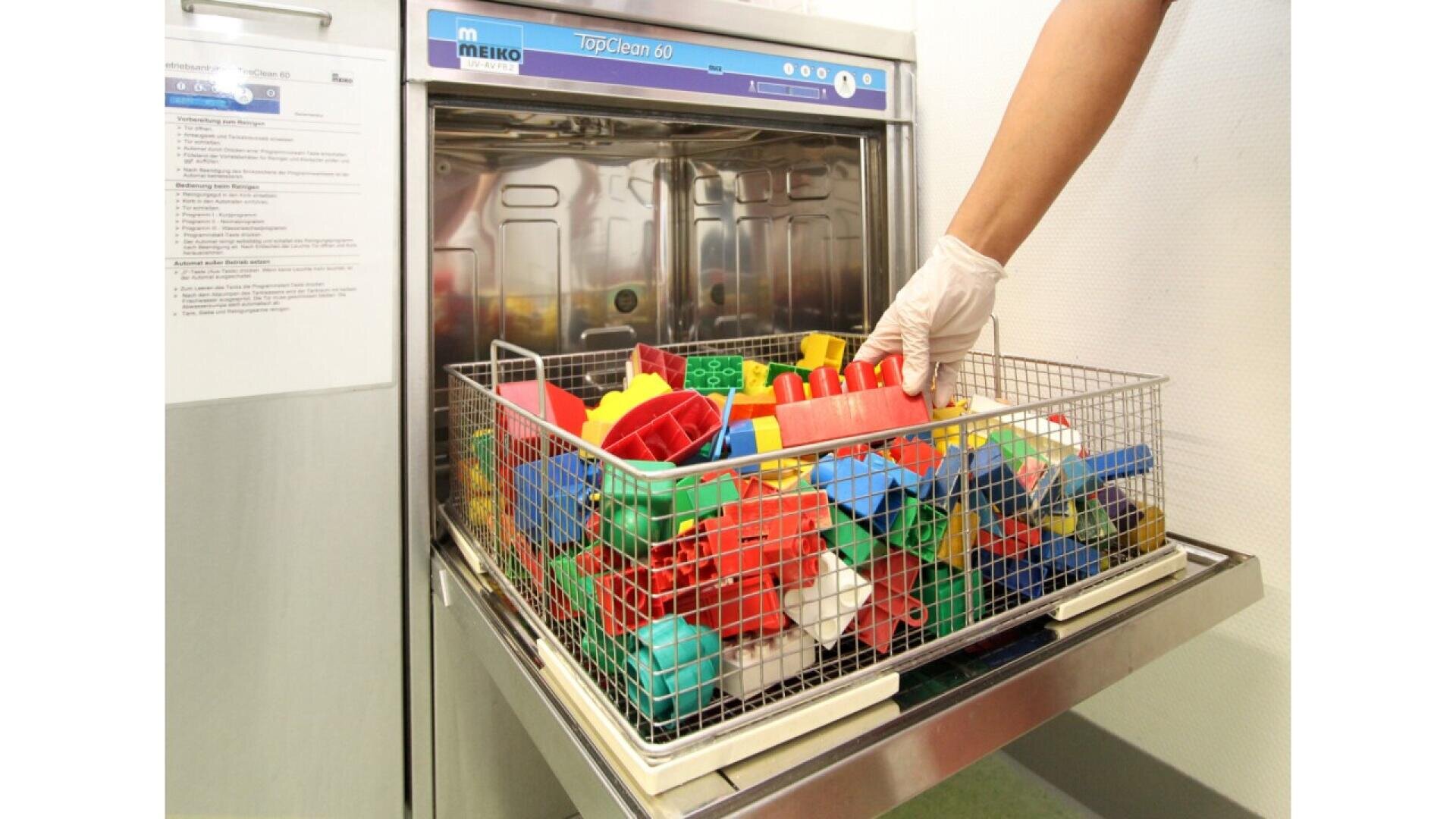 Une main gantée place un panier métallique rempli de blocs de construction en plastique colorés dans un lave-vaisselle industriel.
