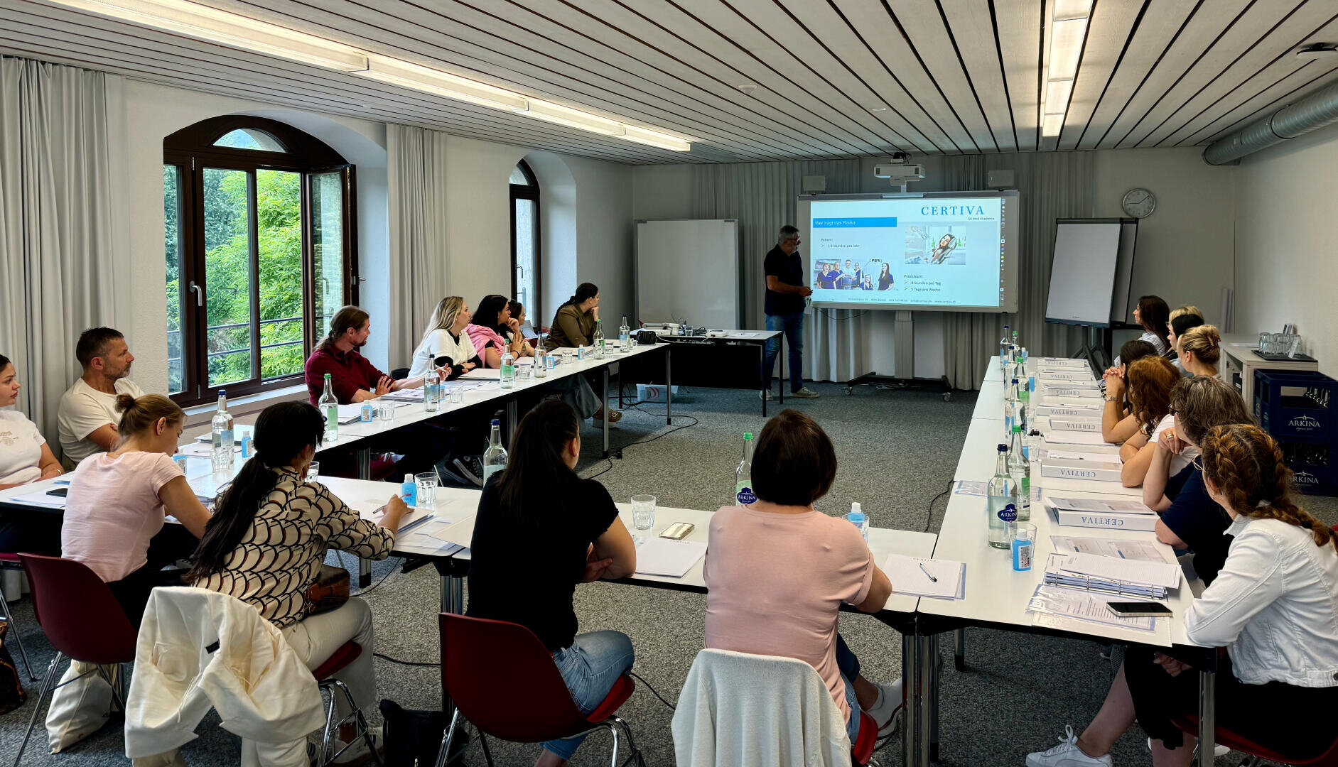 Un groupe de personnes est assis à des tables disposées en forme de U, regardant attentivement un présentateur à l'avant d'une salle de réunion lumineuse avec de grandes fenêtres. Le présentateur fait un geste vers une diapositive projetée sur un écran.