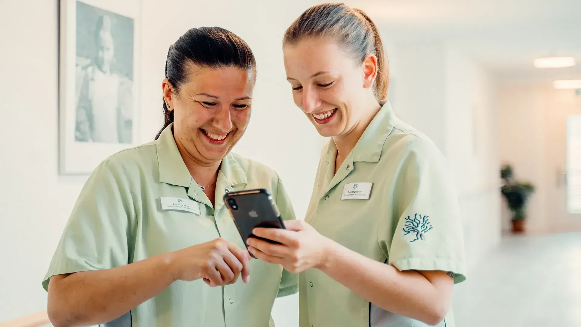 Zwei Mitarbeiter des Gesundheitswesens in hellgrünen Uniformen lächeln und schauen gemeinsam auf ein Smartphone in einem hellen Flur; sie wirken glücklich und engagiert. An der Wand hinter ihnen hängt ein gerahmtes Foto.